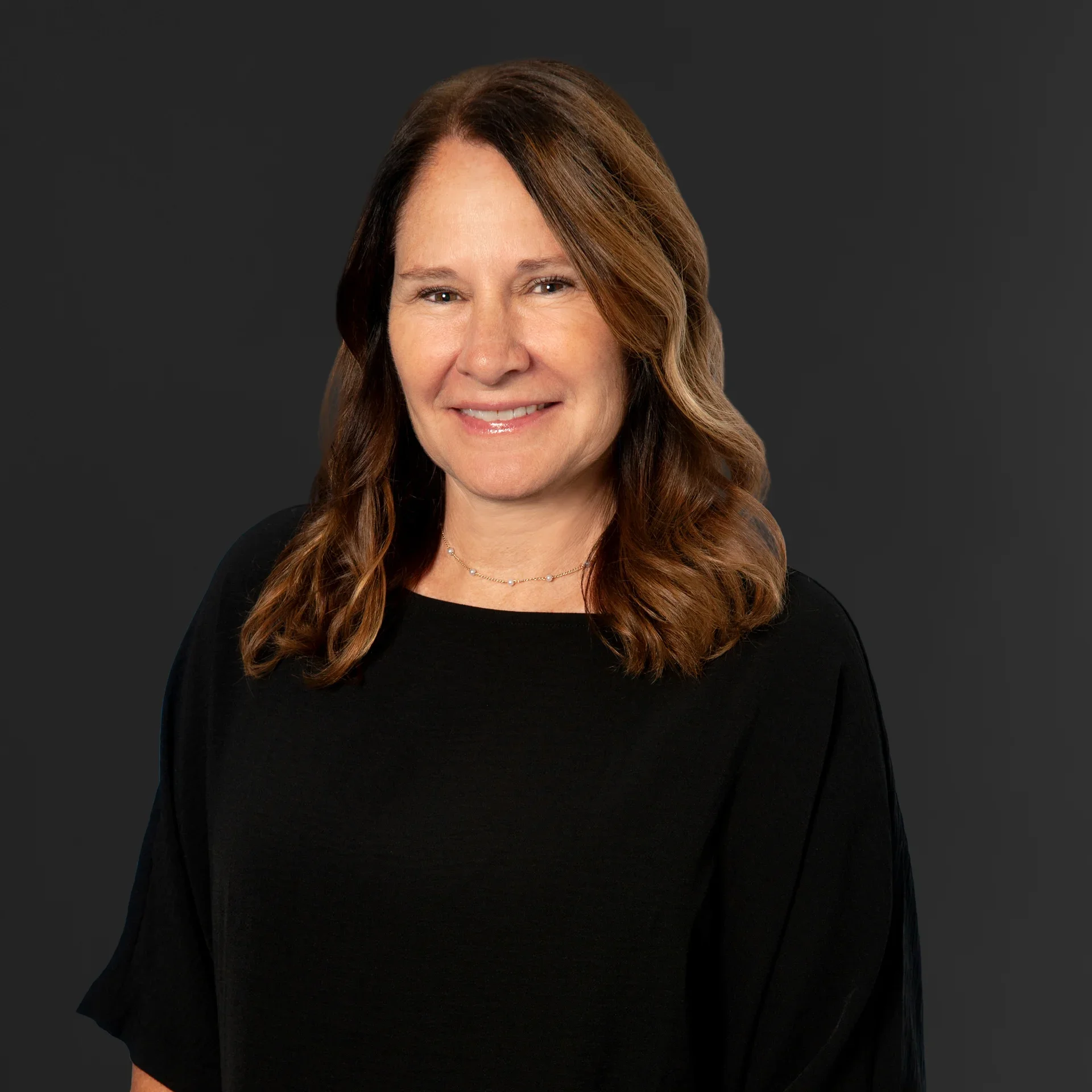 Headshot of a woman with long wavy brown hair, wearing a black top, smiling against a dark background.