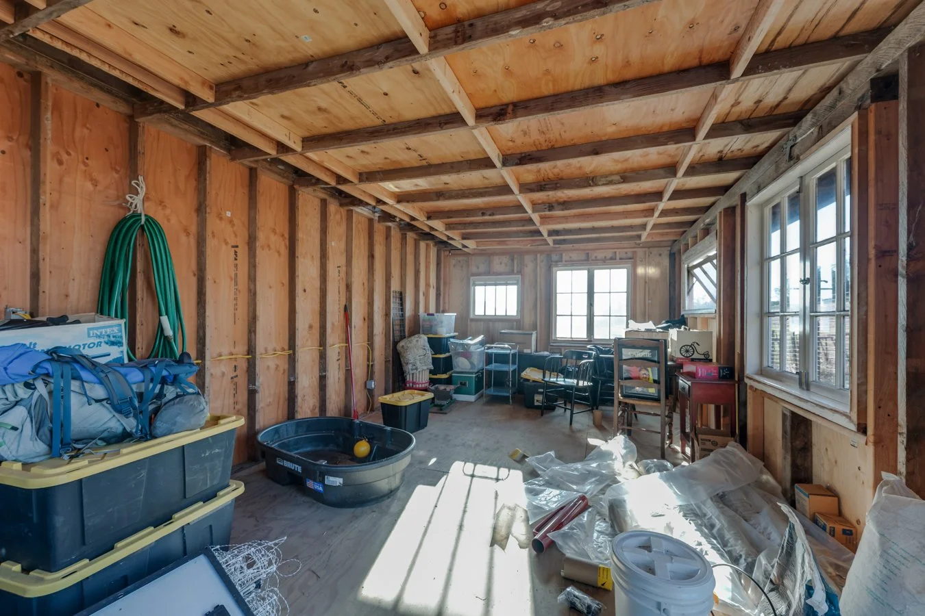 Unfinished room with exposed wooden framing, cluttered with storage bins, tools, and miscellaneous items, with windows allowing natural light.
