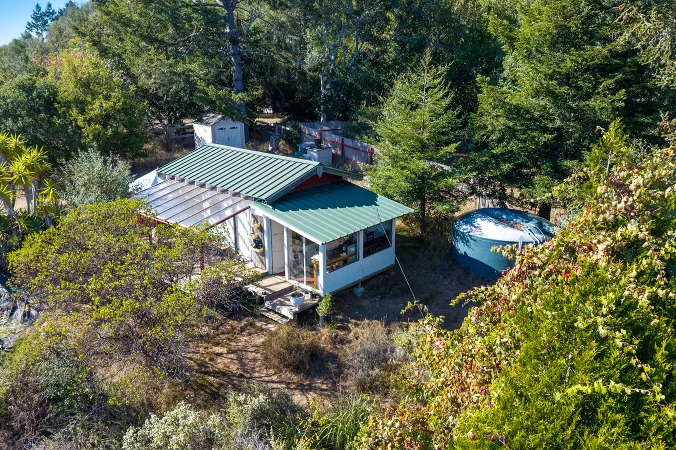 Aerial view of a small house with a green metal roof, surrounded by trees and bushes, with a garden and a water tank nearby.
