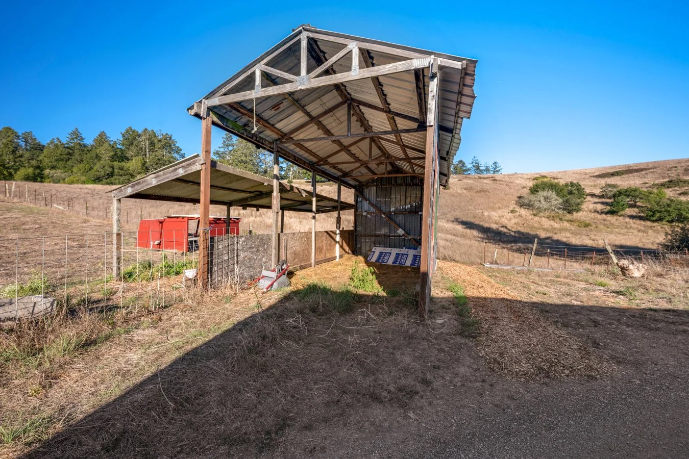 A rural farm scene featuring a partially built shelter with a metal roof and wooden supports, a red storage container in the background, and rolling hills with sparse trees under a clear blue sky.