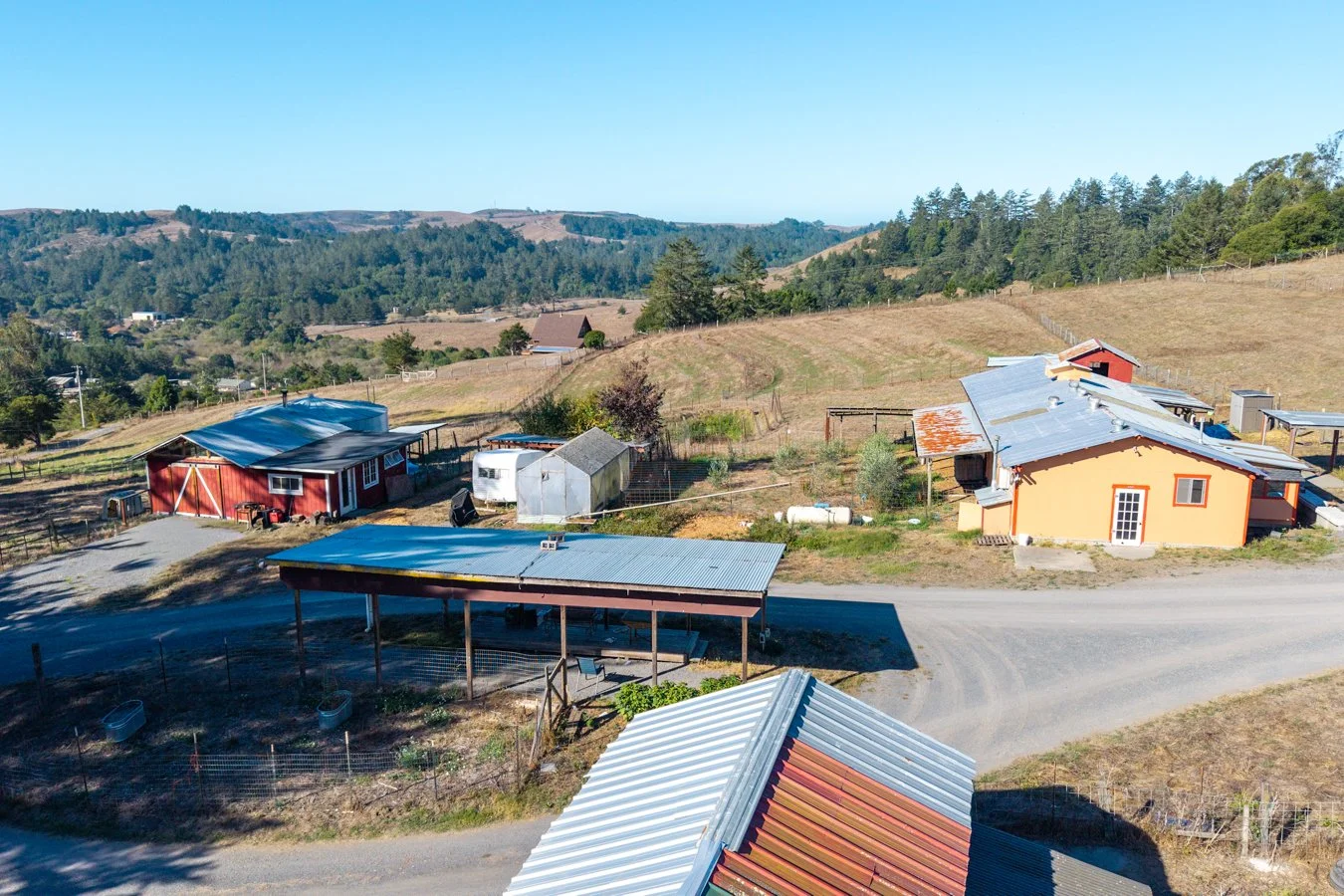 A rural farm scene with several small buildings, greenhouses, fencing, and a dirt road, surrounded by rolling hills and trees under a clear blue sky.