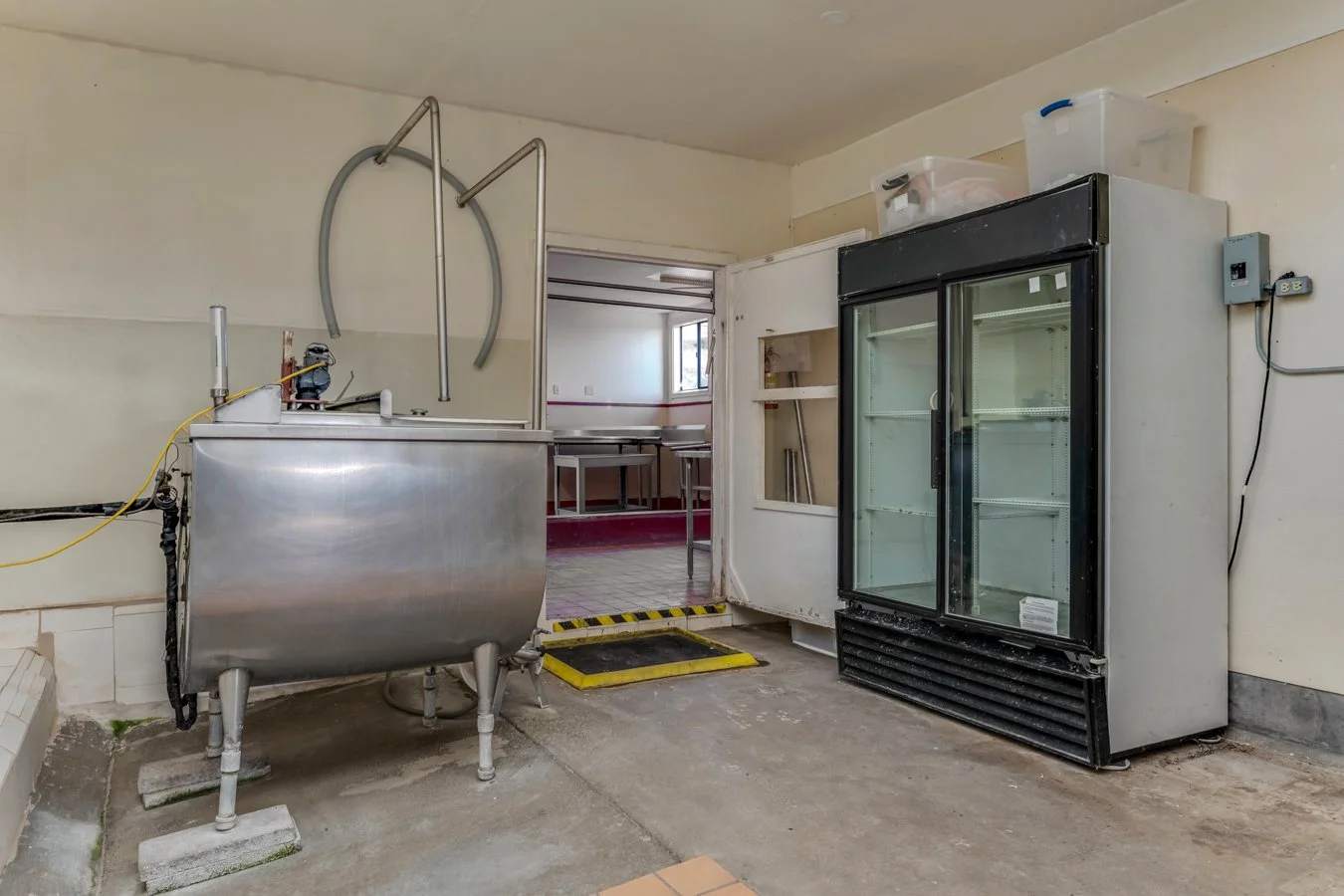 Interior of a commercial kitchen or storage room with a stainless steel sink, a small refrigerator with glass doors, and empty shelving.