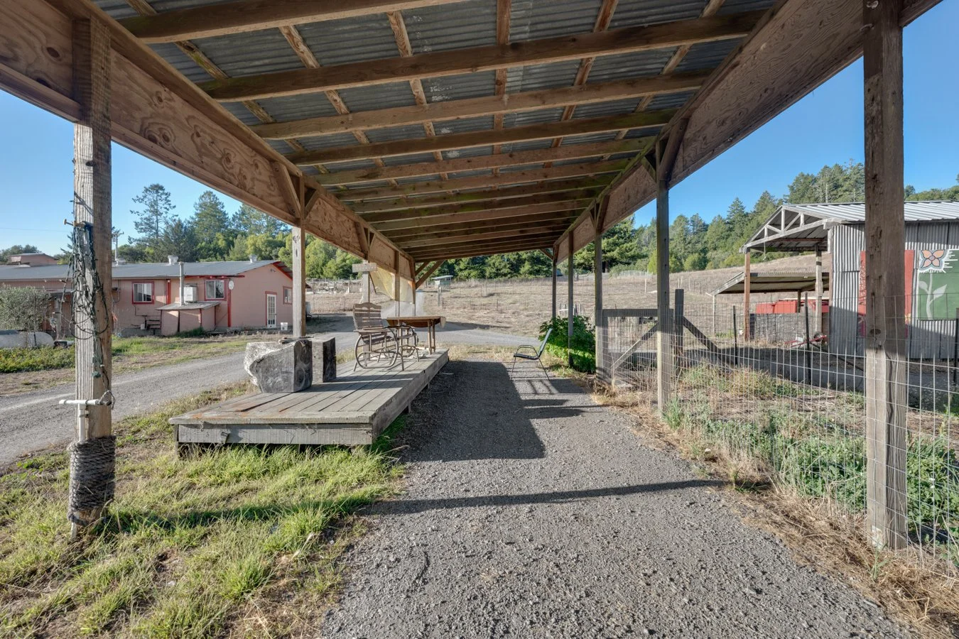 A wooden covered patio with outdoor furniture and a gravel path, with a building and trees in the background.