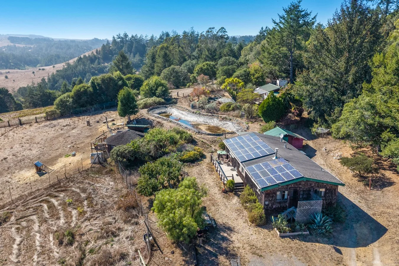 A rural property with a house equipped with solar panels, surrounded by trees and gardens, with a pond and hilly landscape in the background.