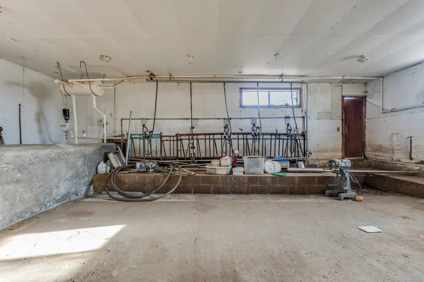 Unfinished basement with plumbing fixtures, hoses, and construction tools, including a saddle tank, pipes, and a door, with an unfinished concrete floor and walls.