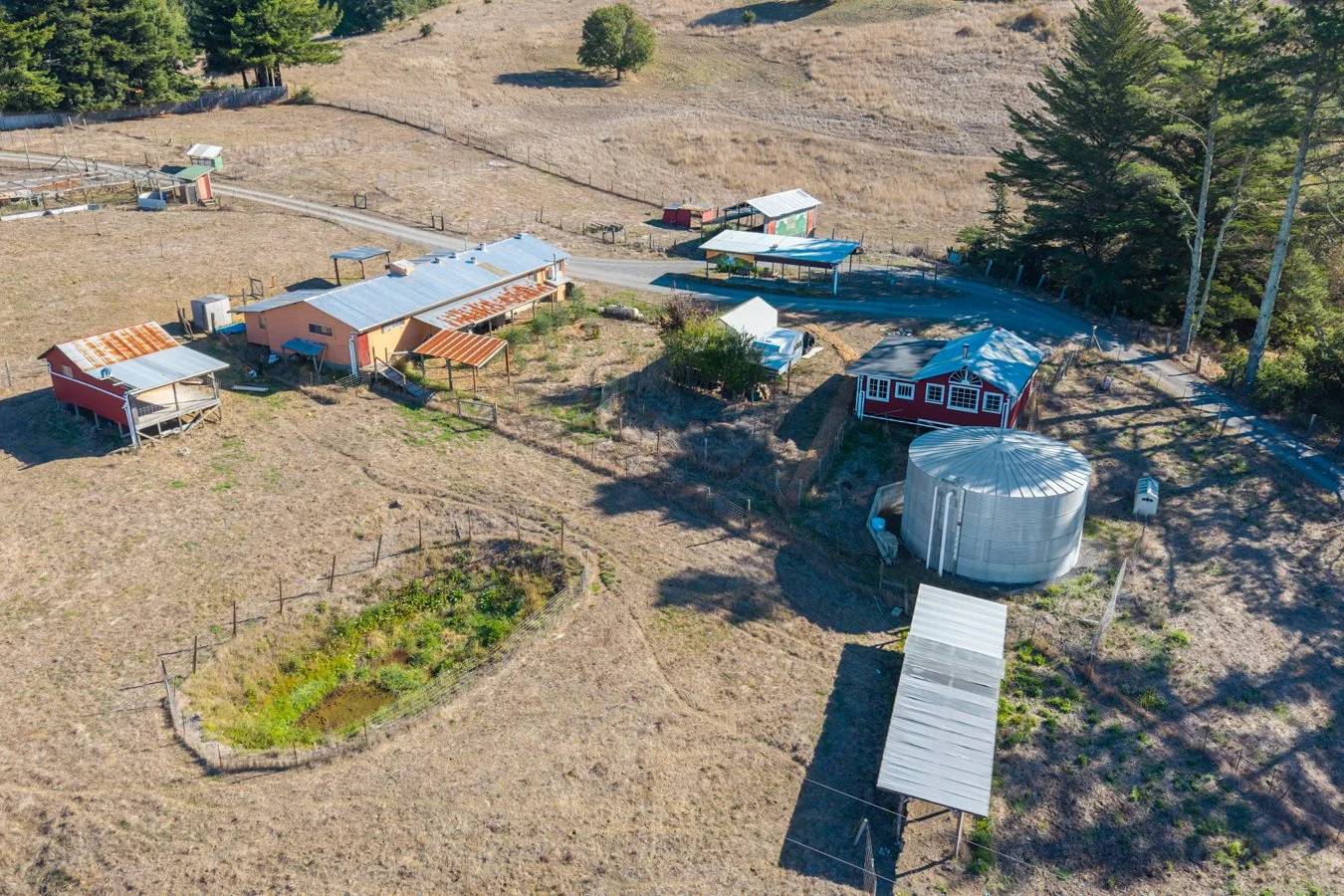 Aerial view of a rural farm with several small buildings, a water tank, and a garden, surrounded by open fields and trees.
