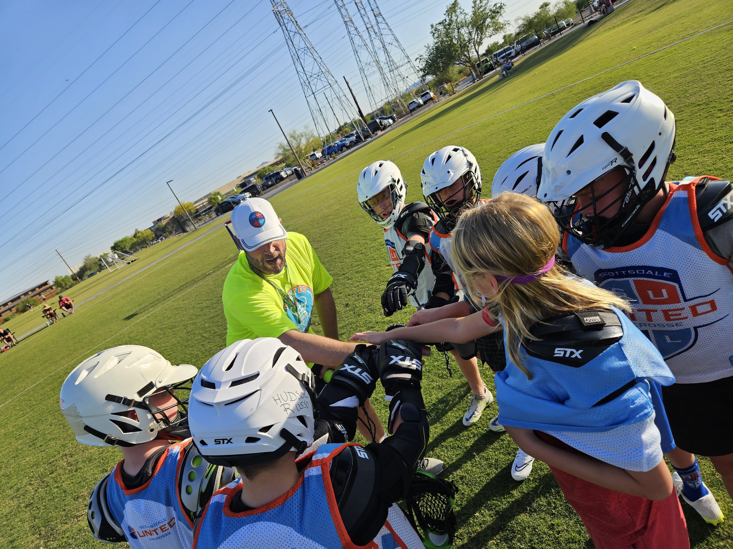 Group of young girls in hockey gear with helmets and jerseys gathered in a circle outside on the field, possibly receiving instructions or encouragement from an adult coach.