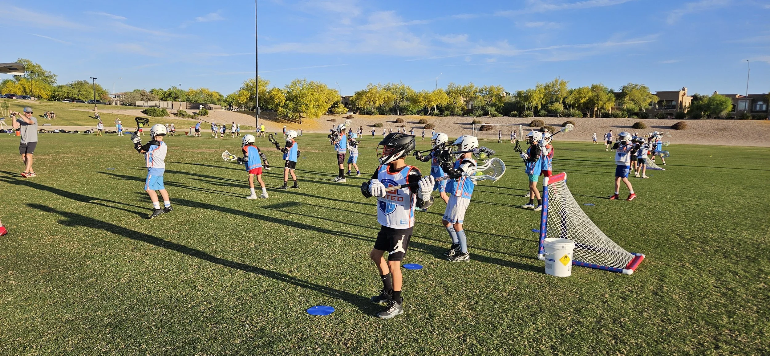 Young children practicing lacrosse on a field with an adult coach overseeing. They are wearing helmets, gloves, and lacrosse gear, with small goals and cones marking the practice area, under a blue sky with some clouds.