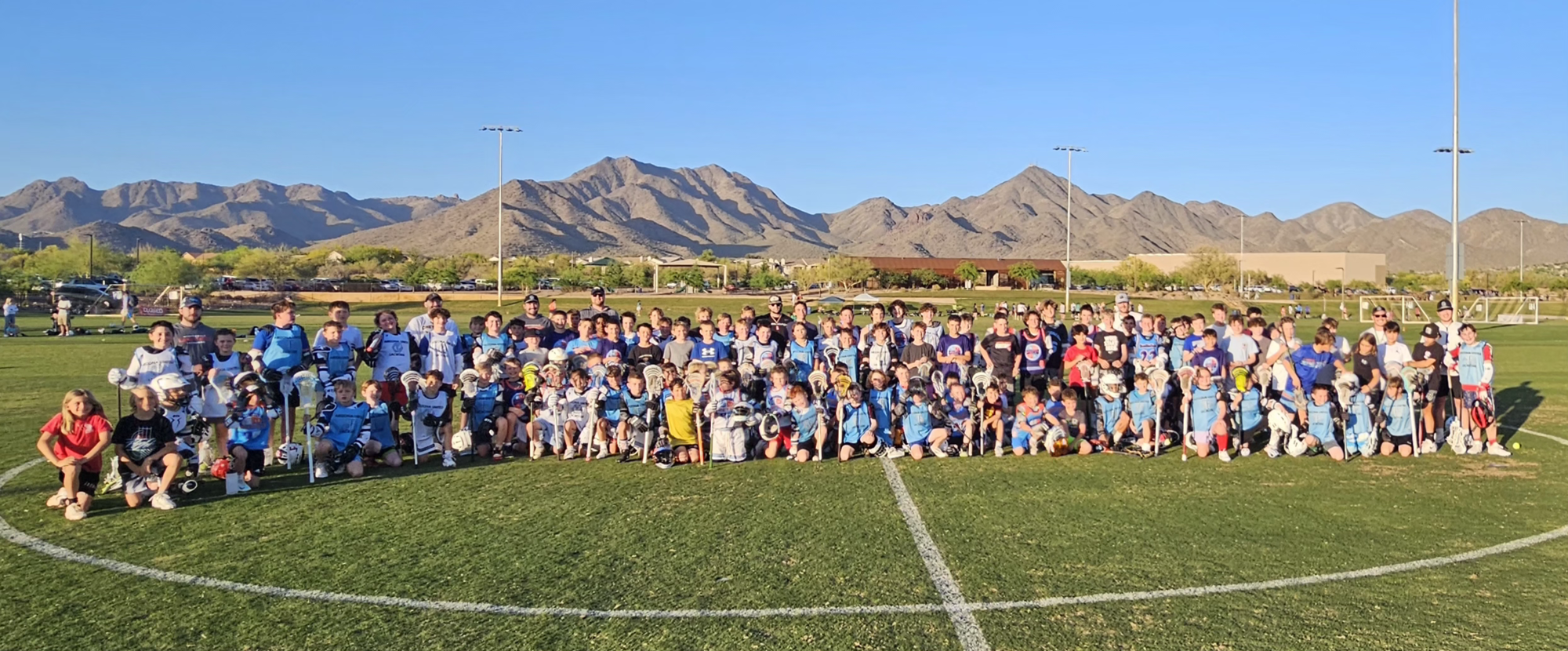 Group of young lacrosse players and coaches posing on a football field with mountains in the background.