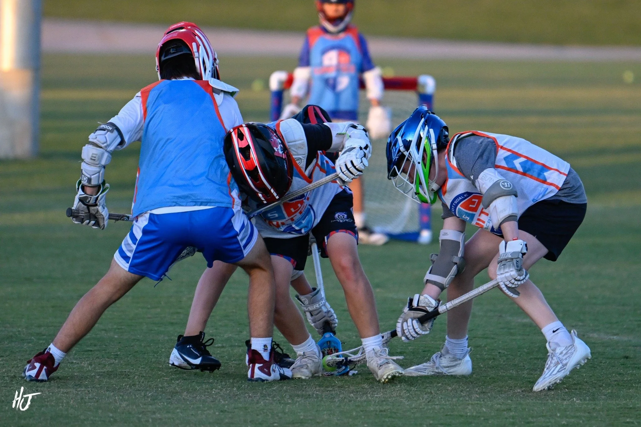 Lacrosse players in helmets and jerseys engaged in a game on a grassy field, with one player appearing to be down on the ground and others involved in a close play.