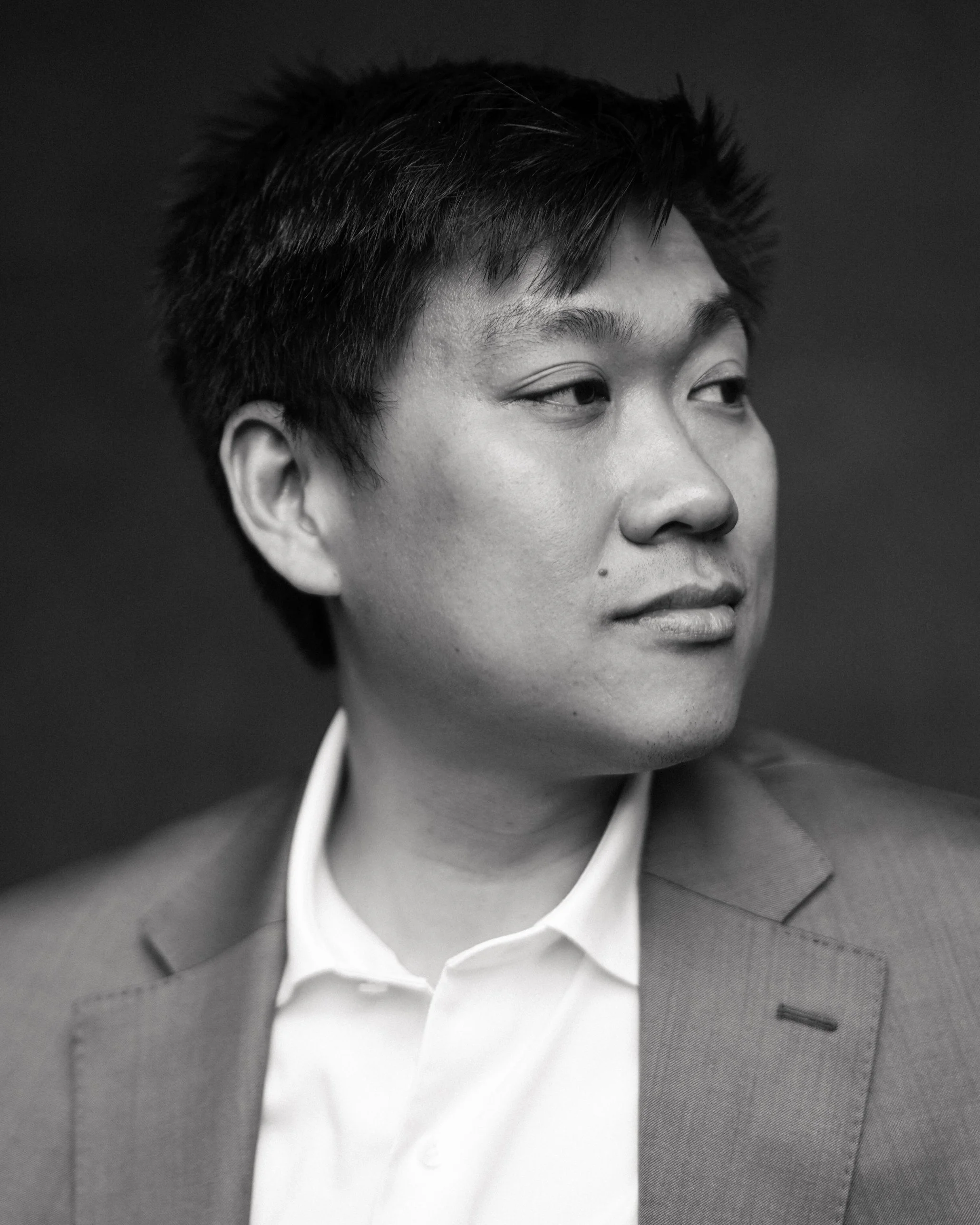 Close-up black-and-white photo of a young man with short, tousled hair wearing a suit jacket and a collared shirt, looking to the side against a plain dark background.