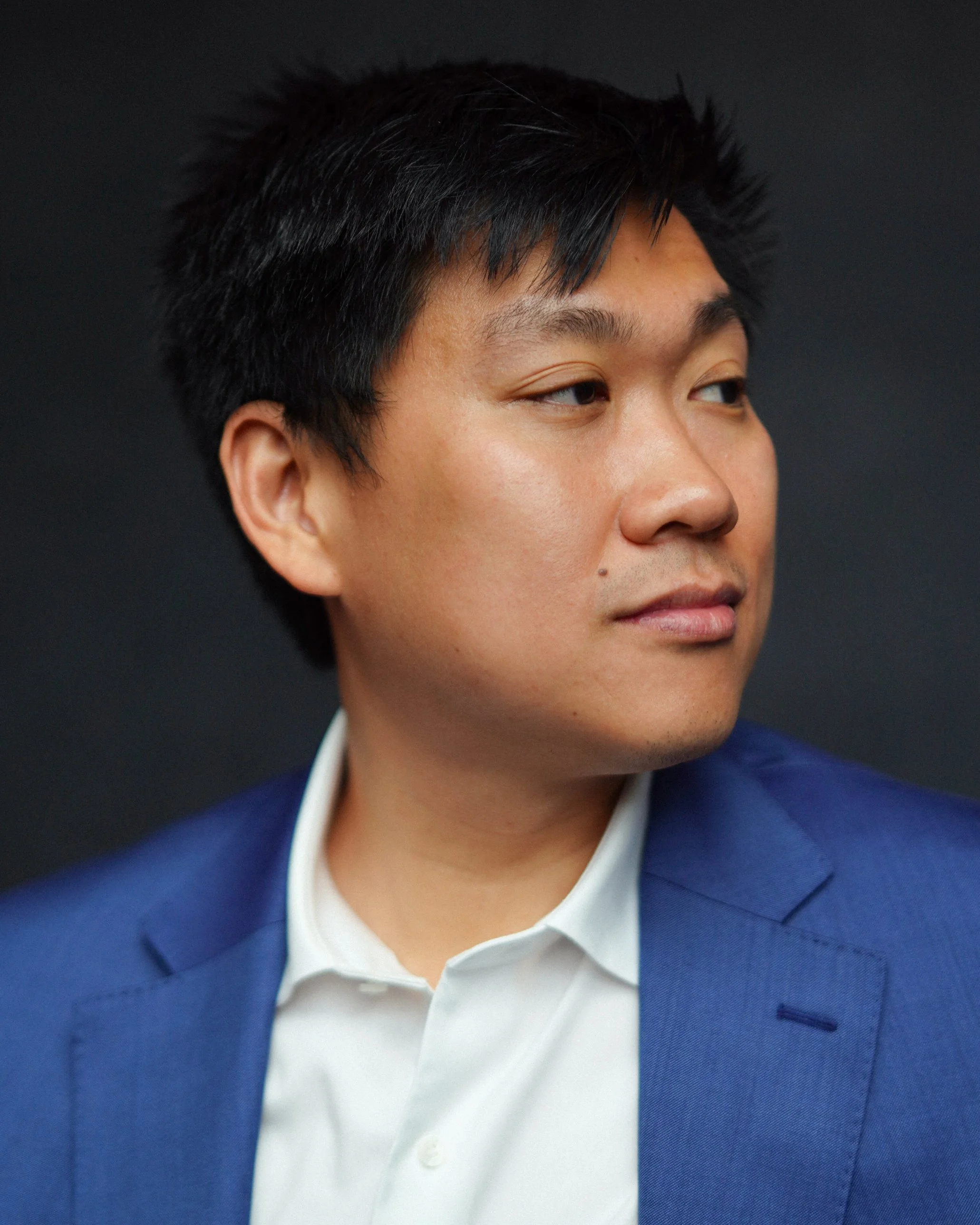 Close-up of a man with black hair in a navy suit and white shirt against a dark background.
