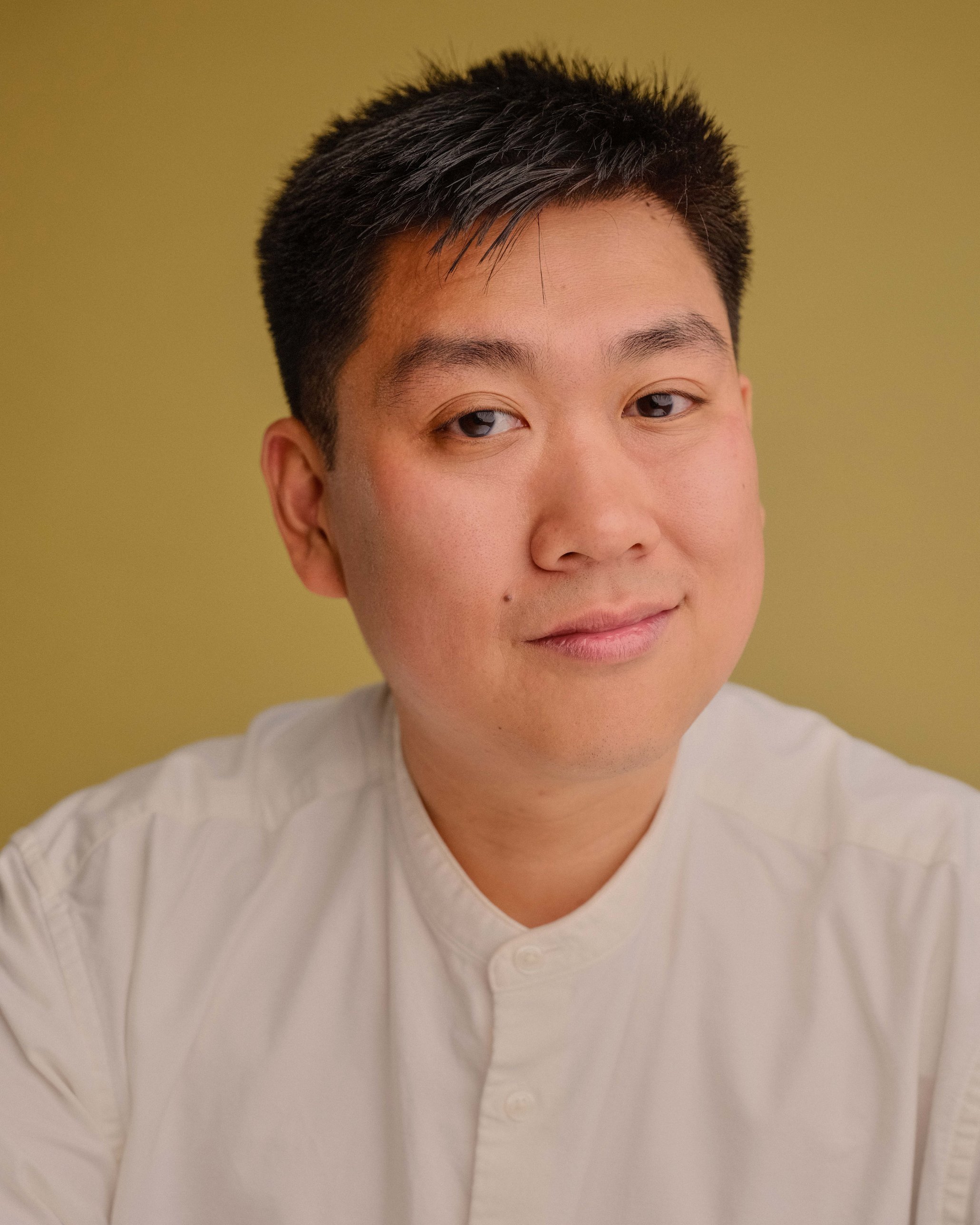 Close-up of a young man with short black hair, light skin, wearing a white textured shirt, against a plain yellow-green background.