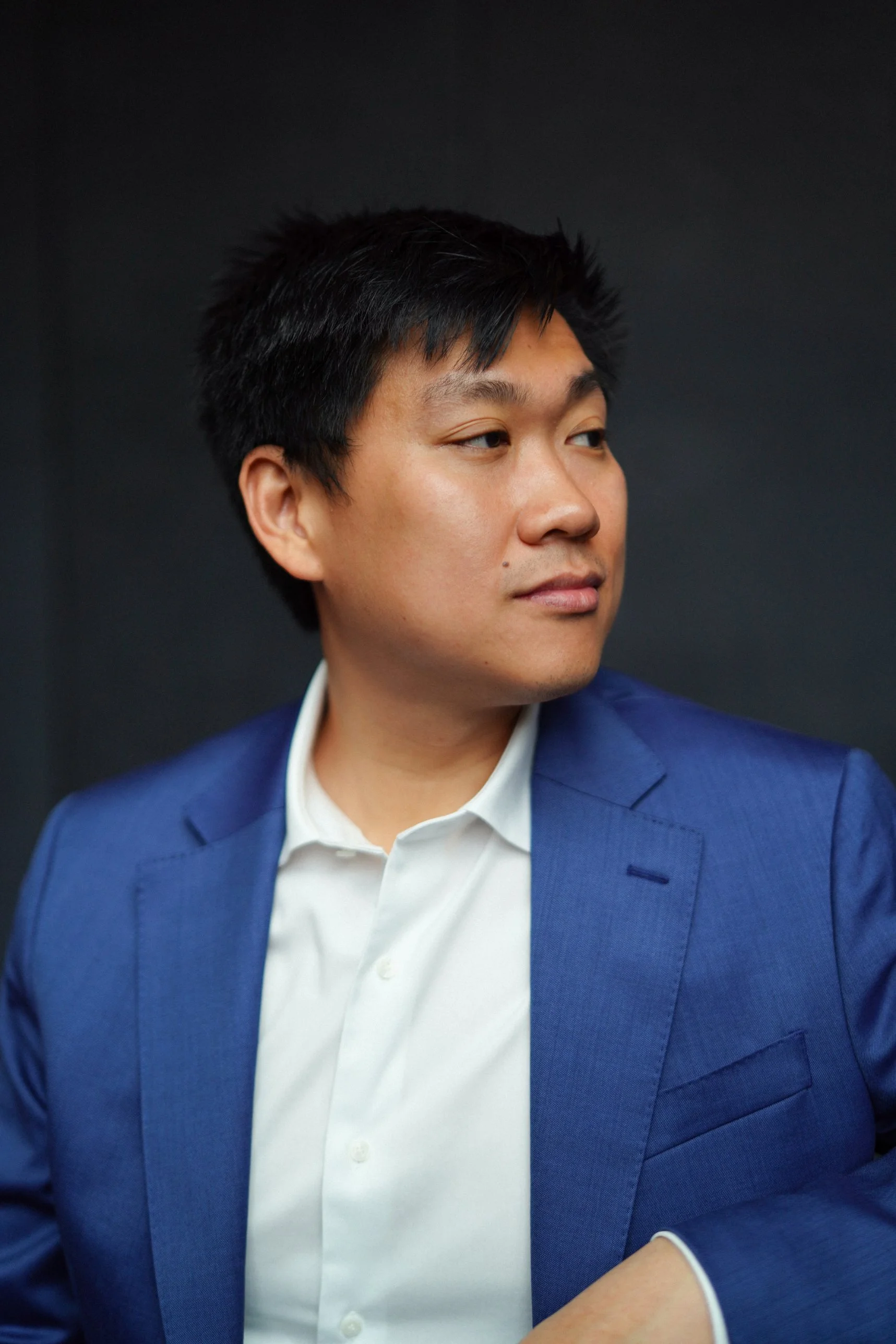 A man wearing a blue suit and white shirt looking to the right against a dark background.