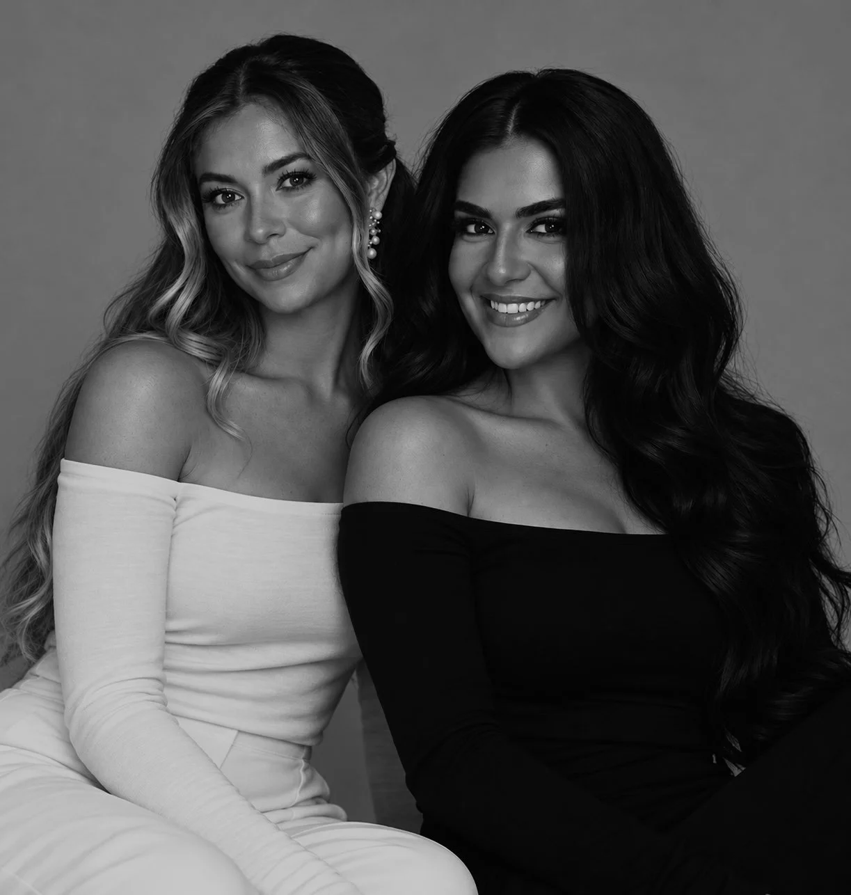 Two smiling women sitting close together, one in a white off-the-shoulder top and the other in a black off-the-shoulder top, against a plain background.