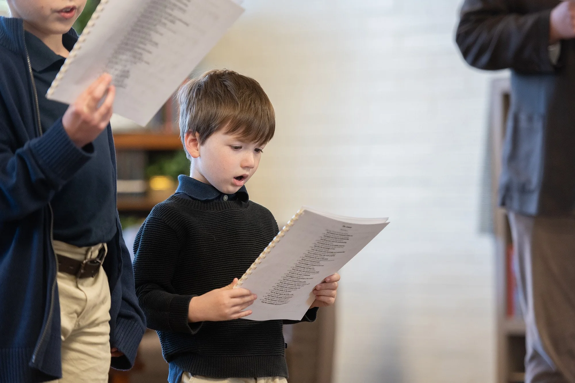 Young boy in a navy sweater reading from a spiral-bound booklet beside other students
