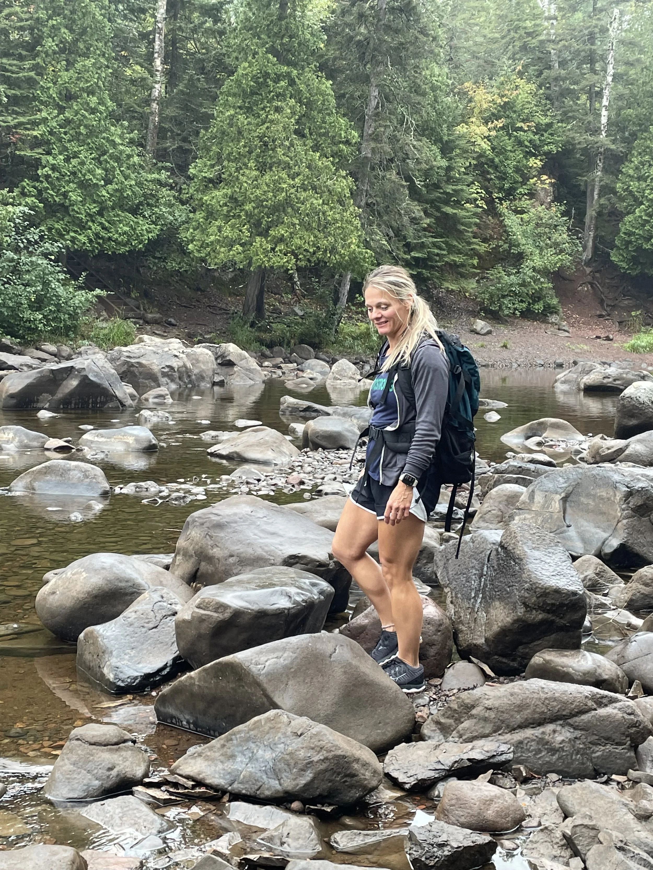 A woman wading through a rocky stream in a forest setting, wearing athletic clothing and a backpack.