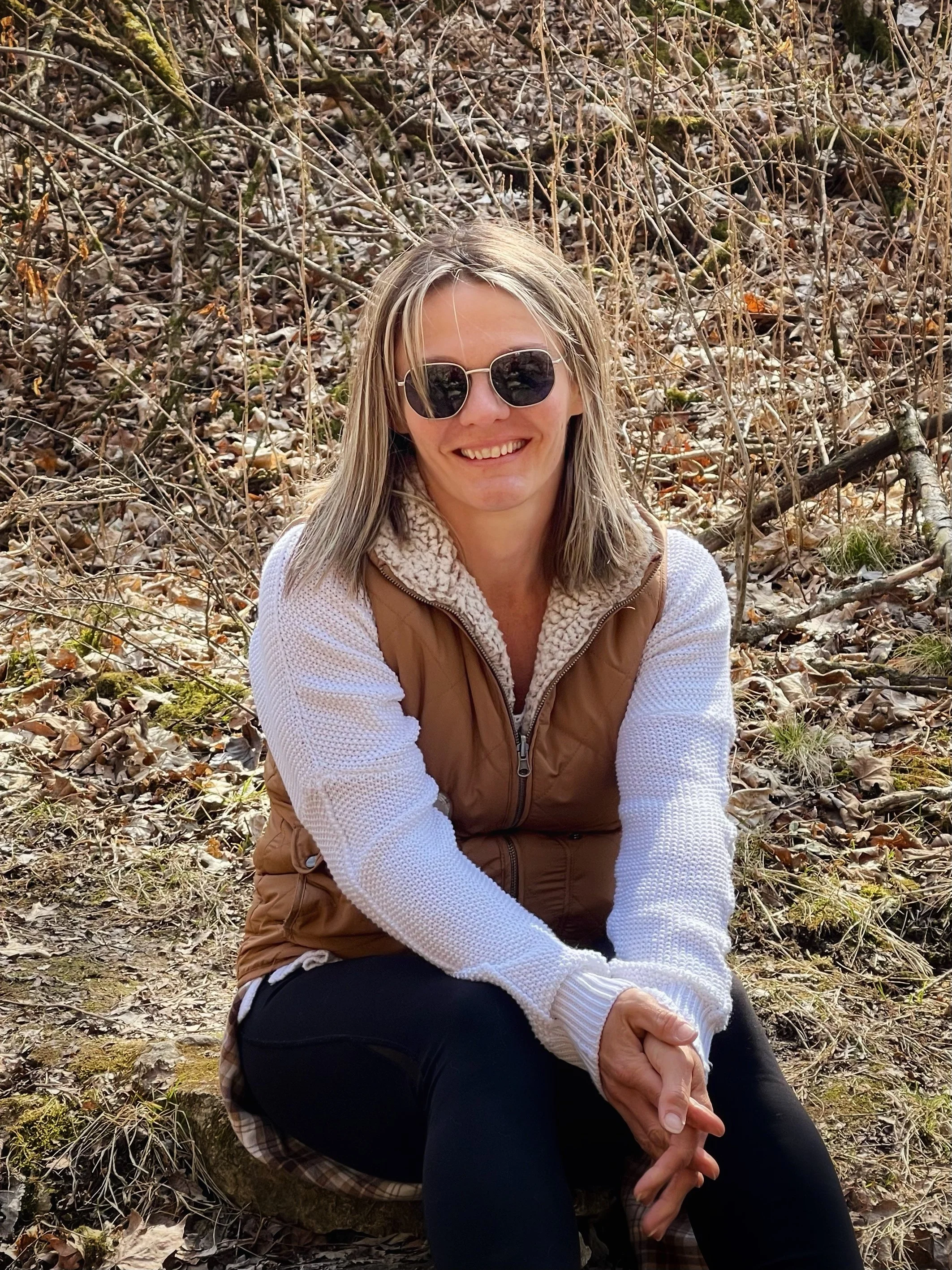 Smiling woman with blonde hair wearing sunglasses, a beige vest, and a white sweater, sitting outdoors on a rock surrounded by fallen leaves and branches.