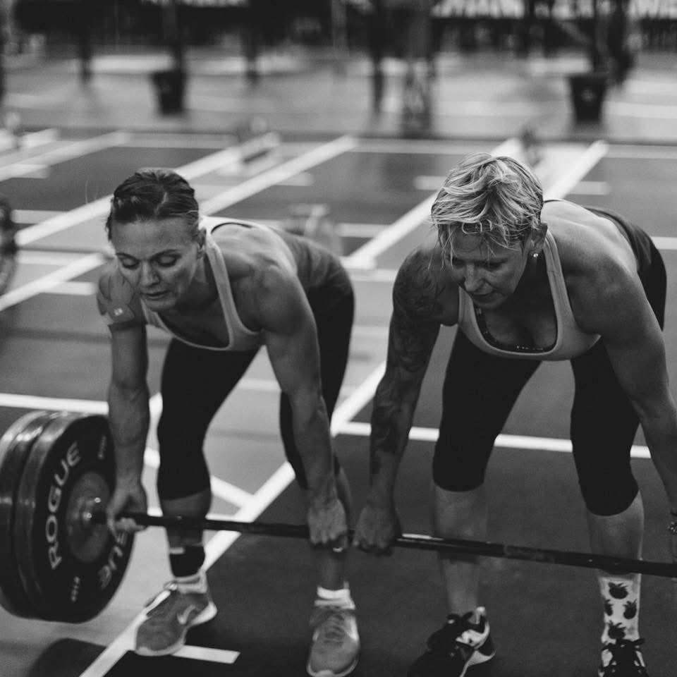 Two women lifting a barbell with weights in a gym, sweating and focused on their workout.