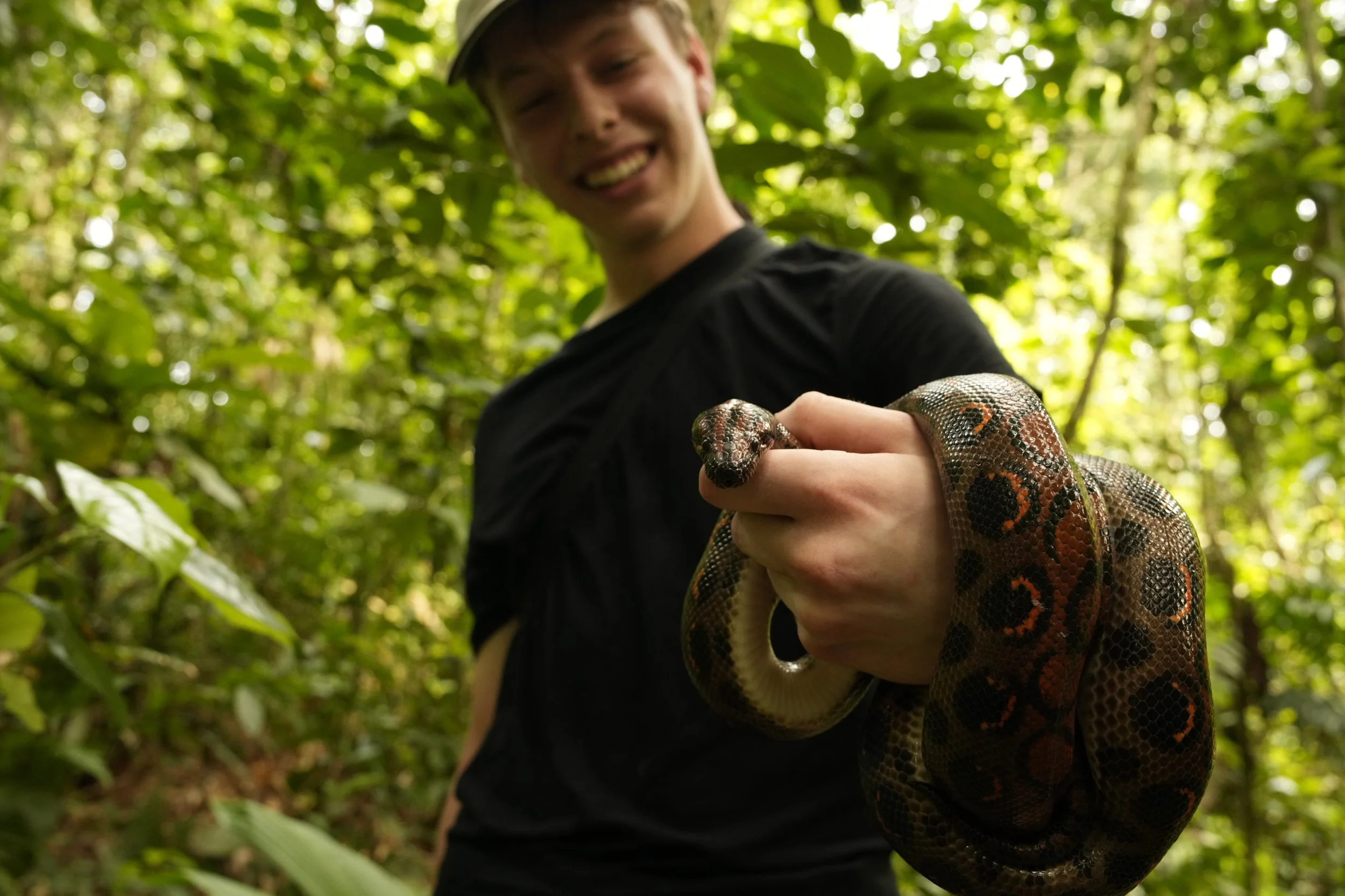 A person in a black shirt holding a snake in a lush green forest, smiling.