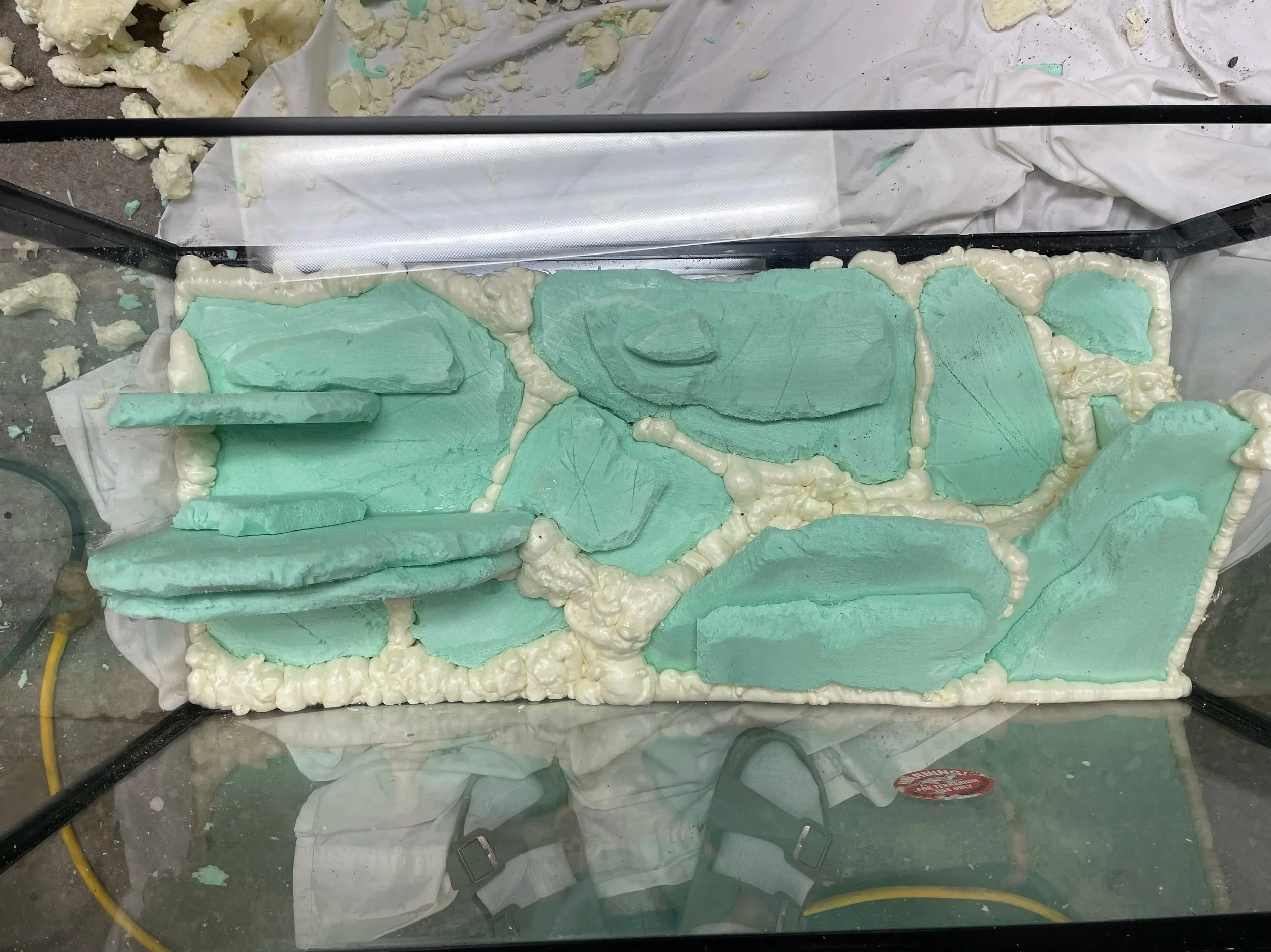 A glass display case with broken foam and foam fragments inside and outside. The case is shattered with cracks and missing pieces, and some foam pieces are on a white cloth underneath.