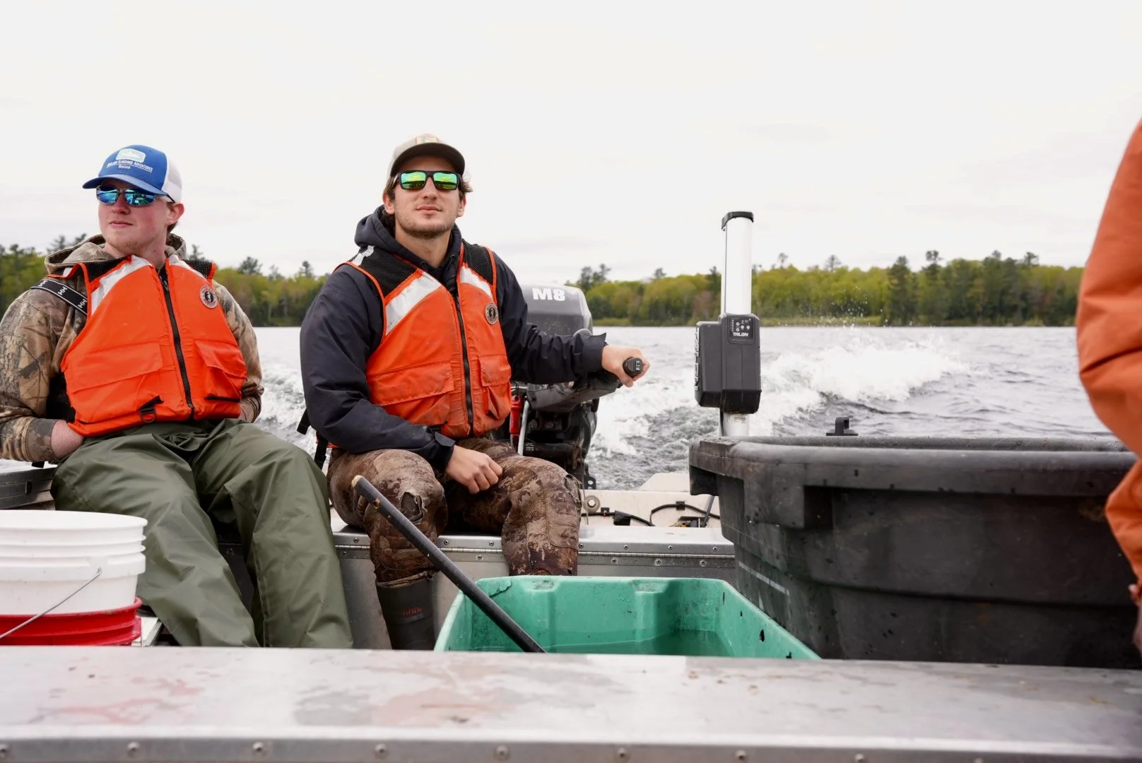 Two men wearing life jackets and camouflage clothing on a boat, with one operating the motor, on a lake with trees in the background.