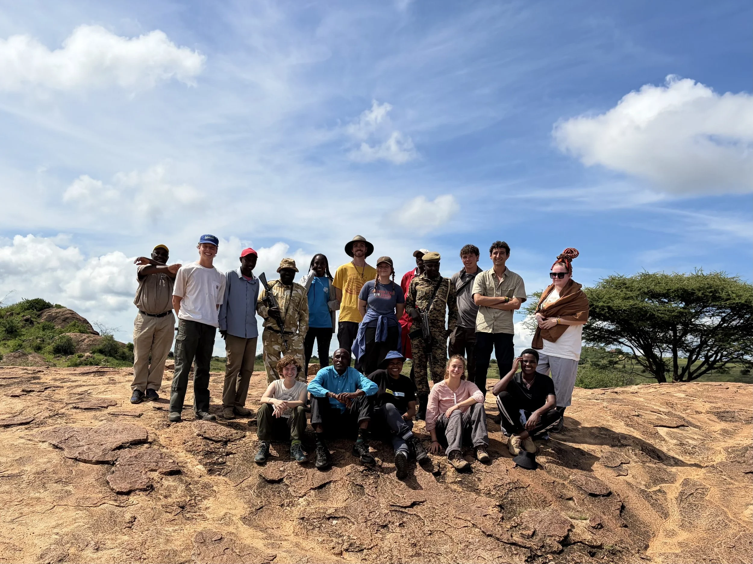 A group of people standing on rocky terrain outdoors under a bright blue sky with scattered clouds.