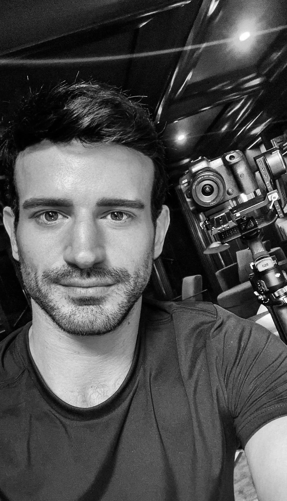 A man with dark hair and stubble taking a selfie with a professional camera mounted on a tripod in a dimly-lit interior.