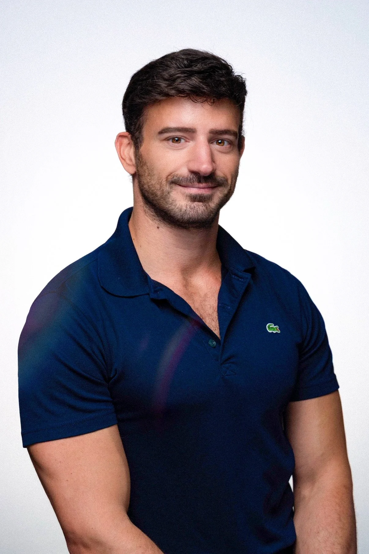Portrait of a man with dark hair and a beard, wearing a navy blue Lacoste polo shirt, standing against a plain white background.