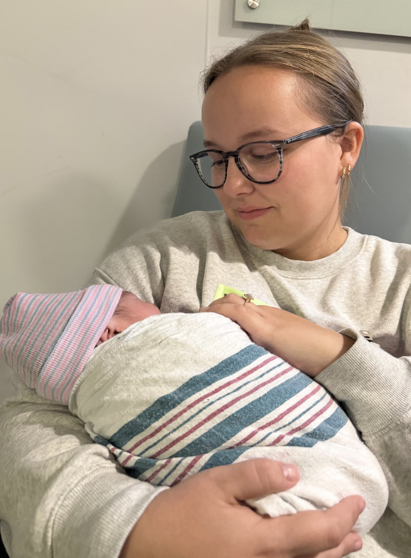 A doula with glasses and earrings holds a newborn baby wrapped in a striped hospital blanket and wearing a pink striped cap, in a hospital room.