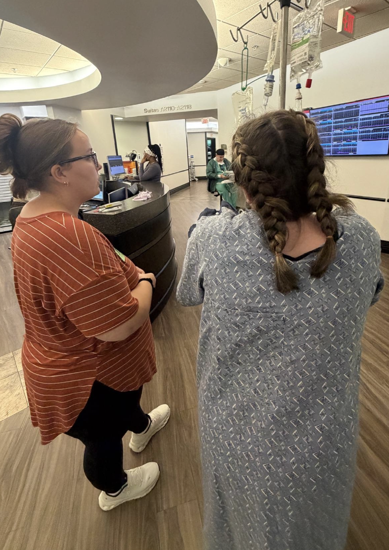 Two women, one wearing an orange striped shirt and glasses is the diula, and the other in a hospital gown with braids, standing in a hospital reception area, talking. In the background, healthcare staff and medical equipment are visible.