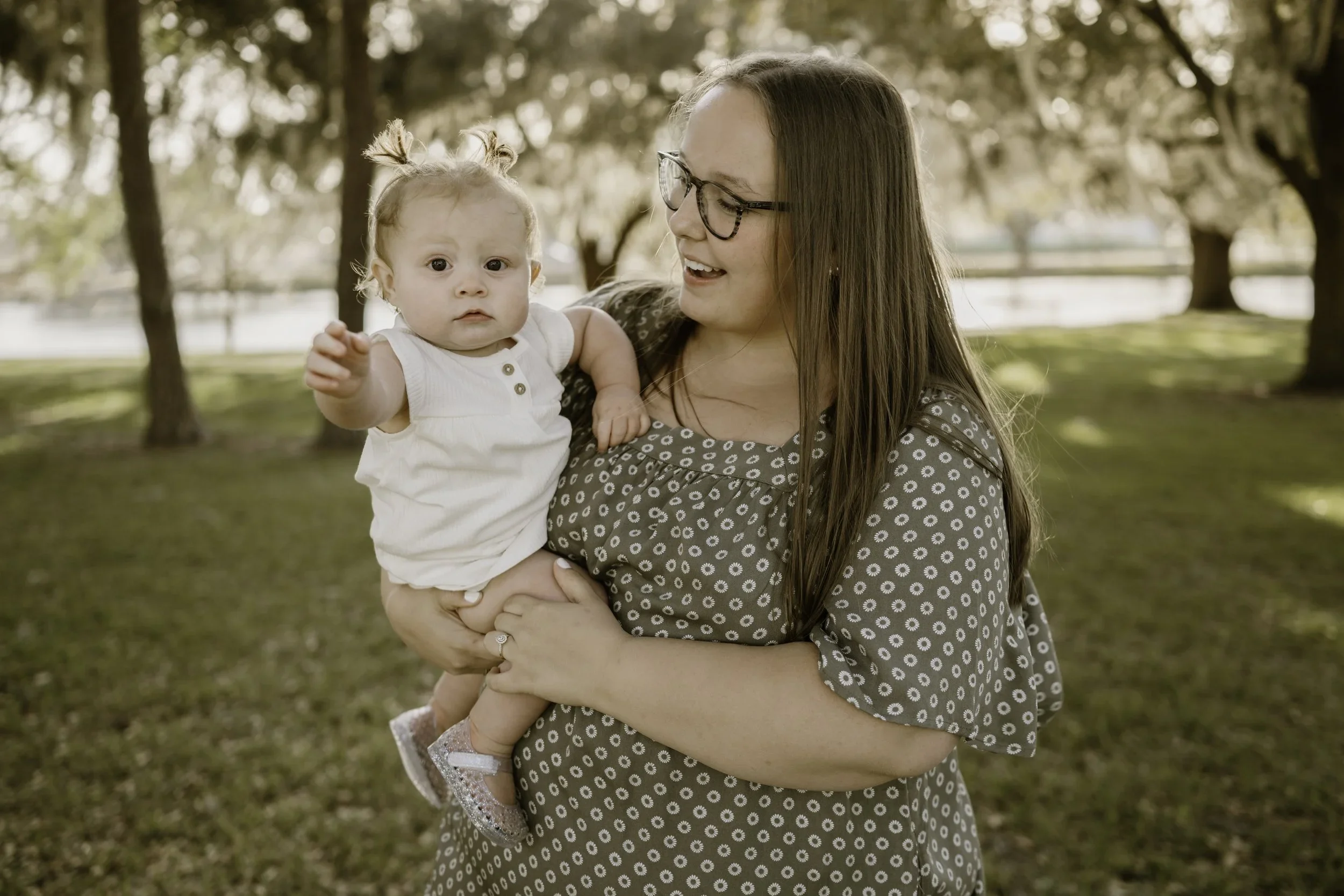 A woman with glasses holding a young girl with two ponytails in an outdoor park with trees and a fence in the background.