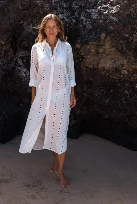 Model in a white eyelet button-front midi shirtdress against dark rocks