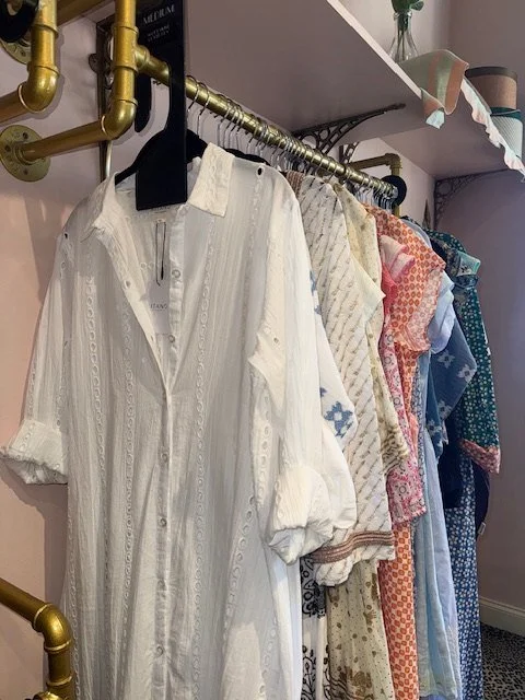 Rack of resort wear in the shop: a white eyelet shirtdress beside colorful printed dresses
