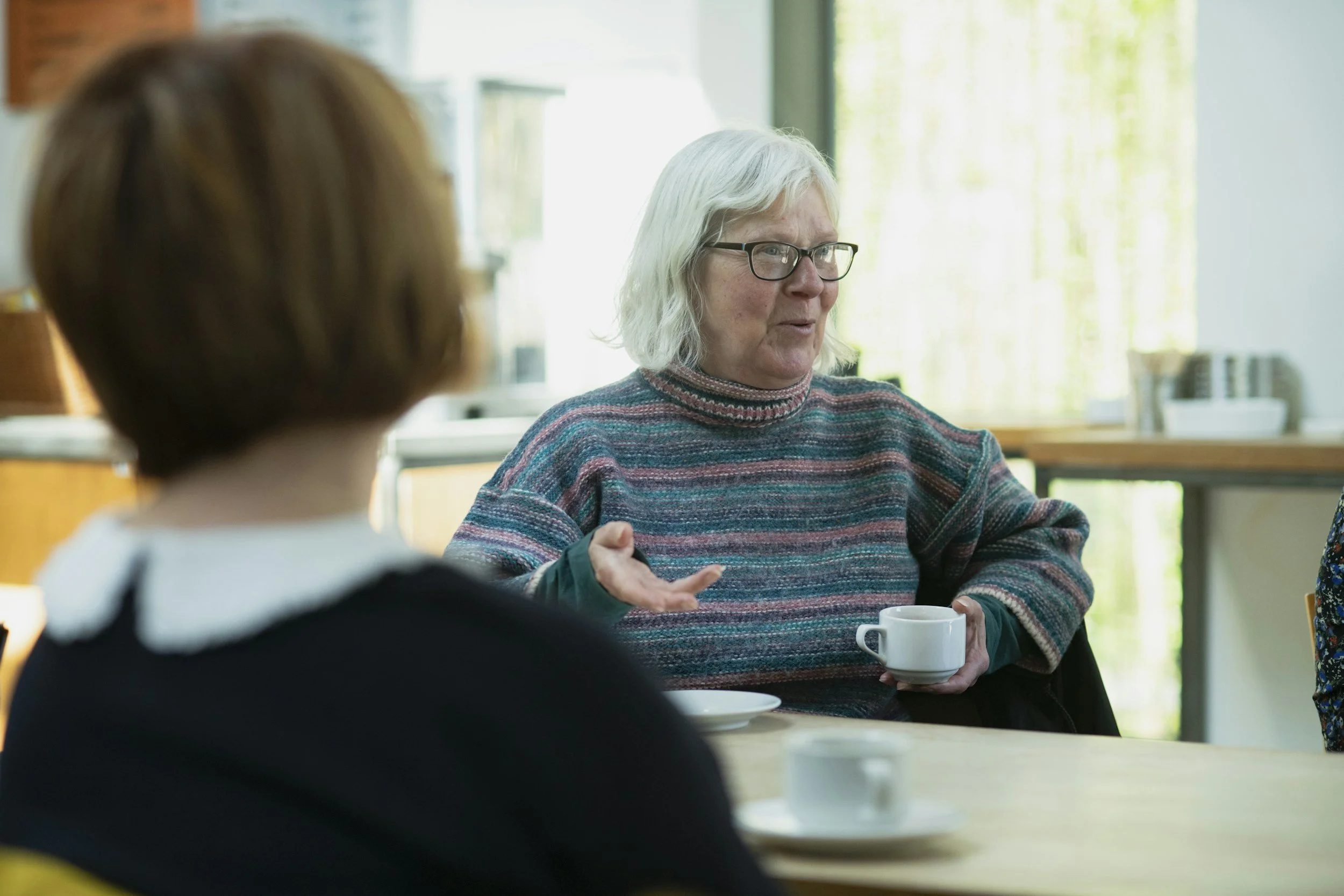 An older woman talking with another person over coffee at a table.