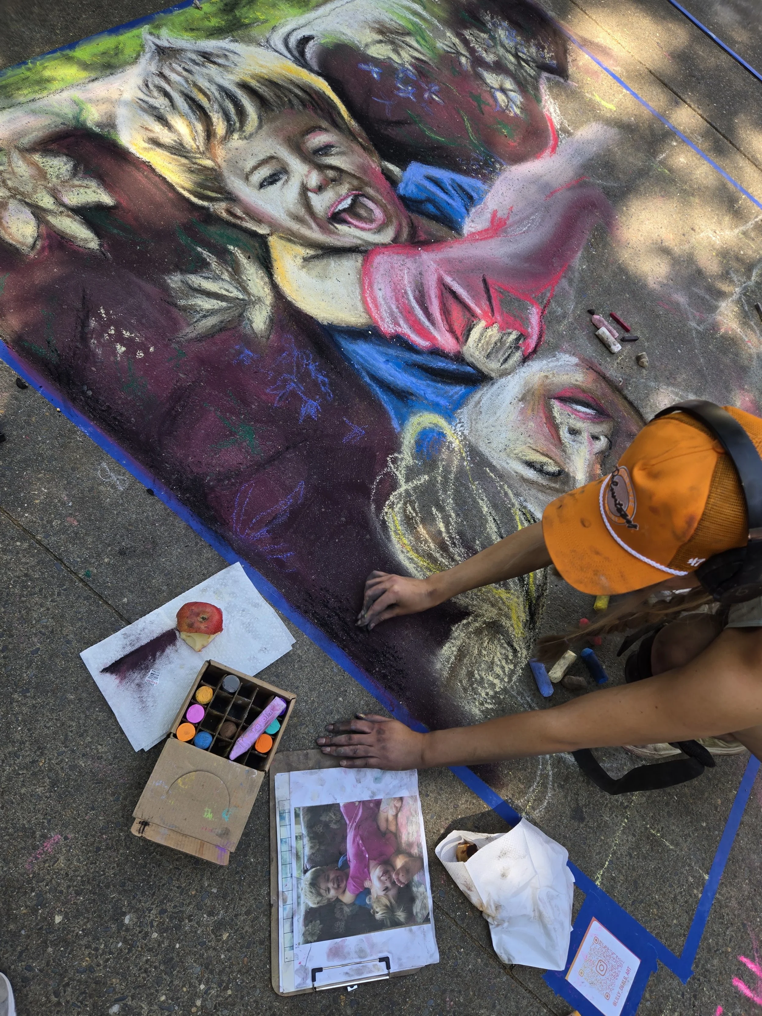 A person creates a colorful chalk art portrait of a smiling young girl with short hair. Art supplies and reference images are nearby on the ground.