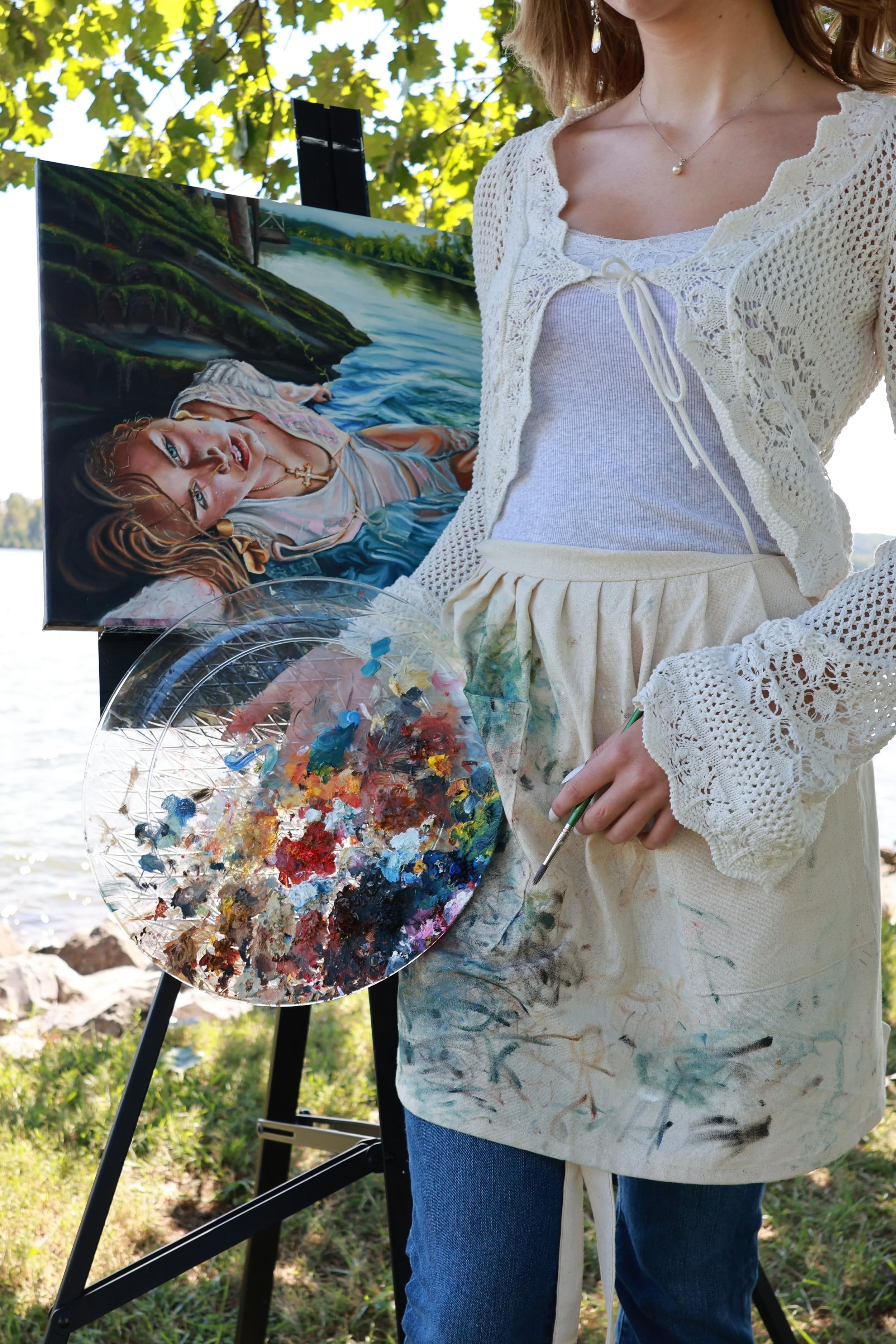 Person outdoors, holding a paint palette with mixed colors, wearing a white lace top and jeans, standing next to an easel with a painting of a woman by a river, trees, and a bridge.