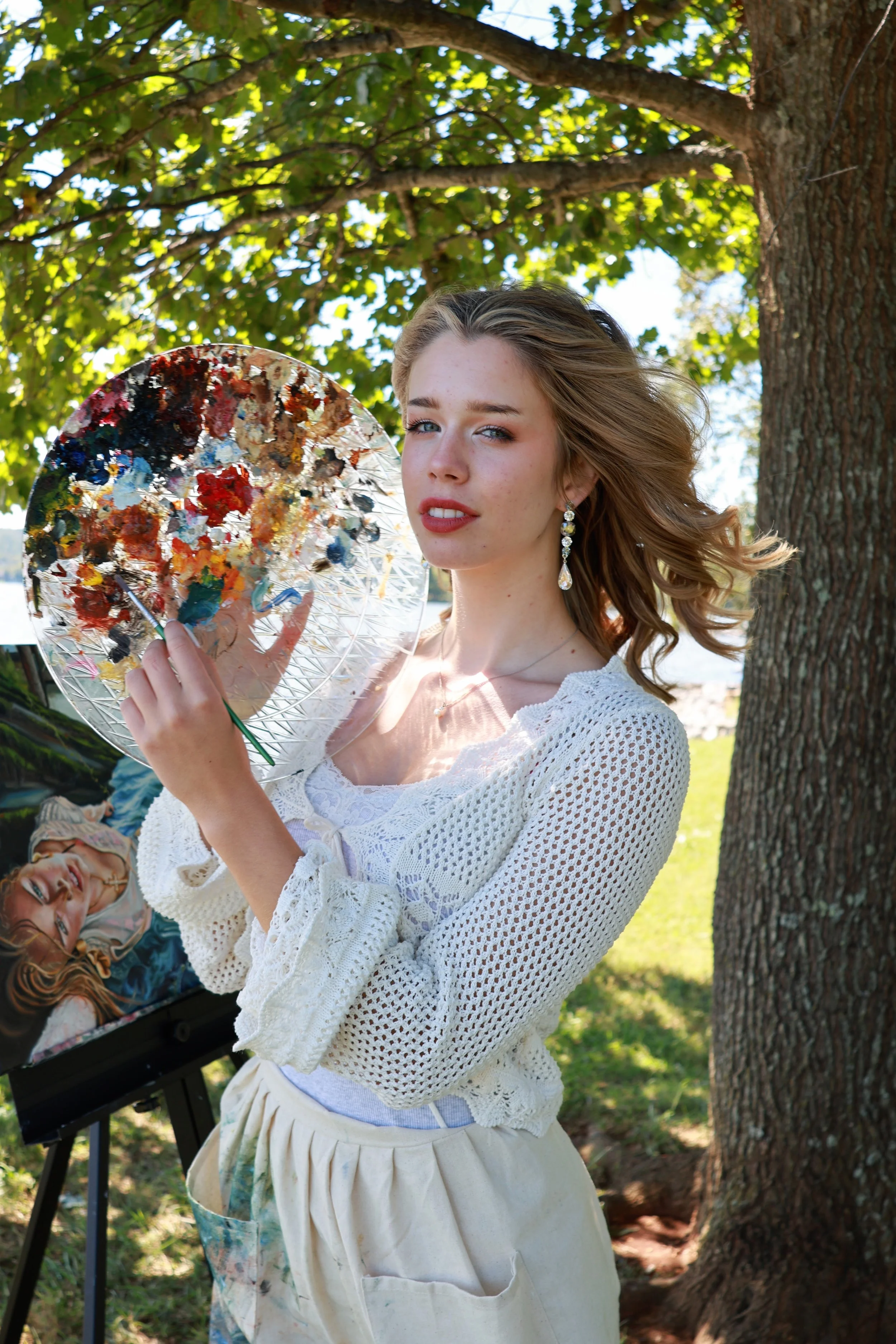 Young woman outdoors holding a painter's palette with various paint colors, standing near a tree with green leaves, wearing a white crocheted top, light-colored skirt, and earrings.