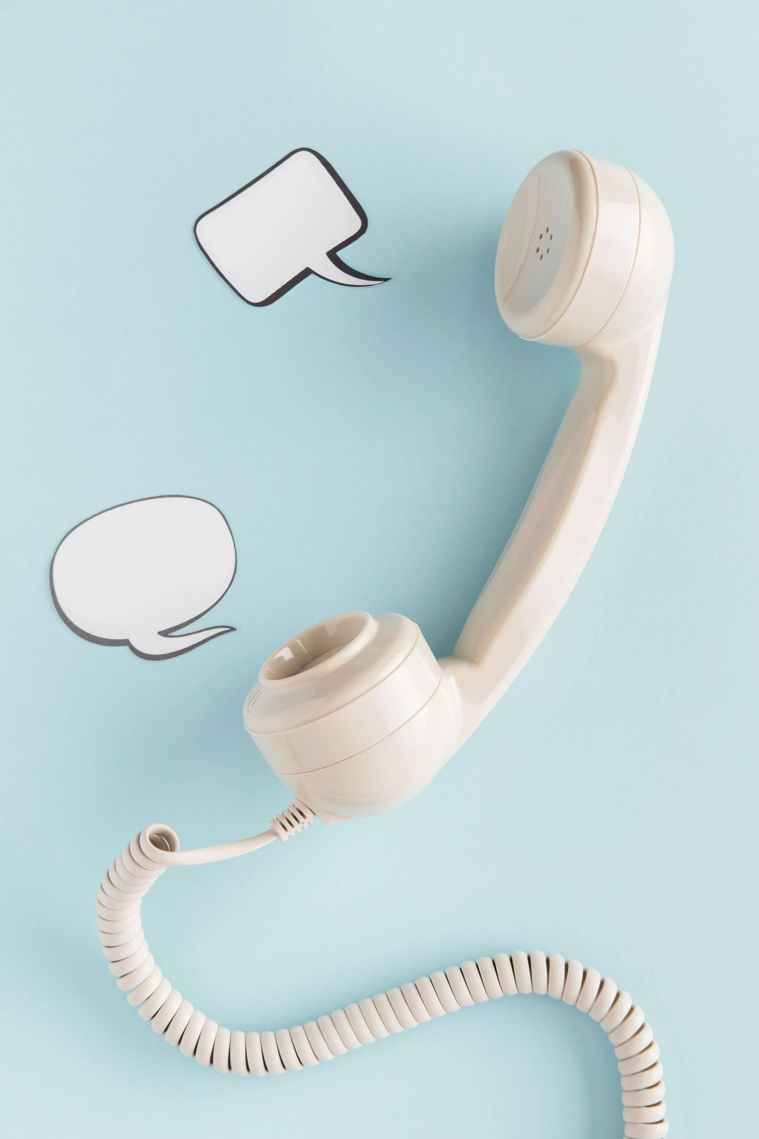 A white telephone receiver connected to a coiled cord is placed on a pastel blue background. Two speech bubbles are illustrated nearby, one above and one below, indicating conversation.