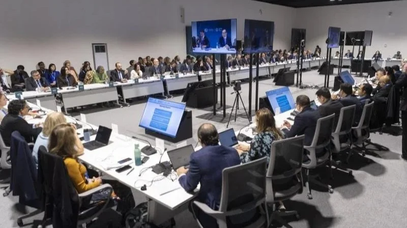 Large conference room with many people seated around a U-shaped table, many using laptops and papers, with large screens displaying the meeting and speakers at the front.