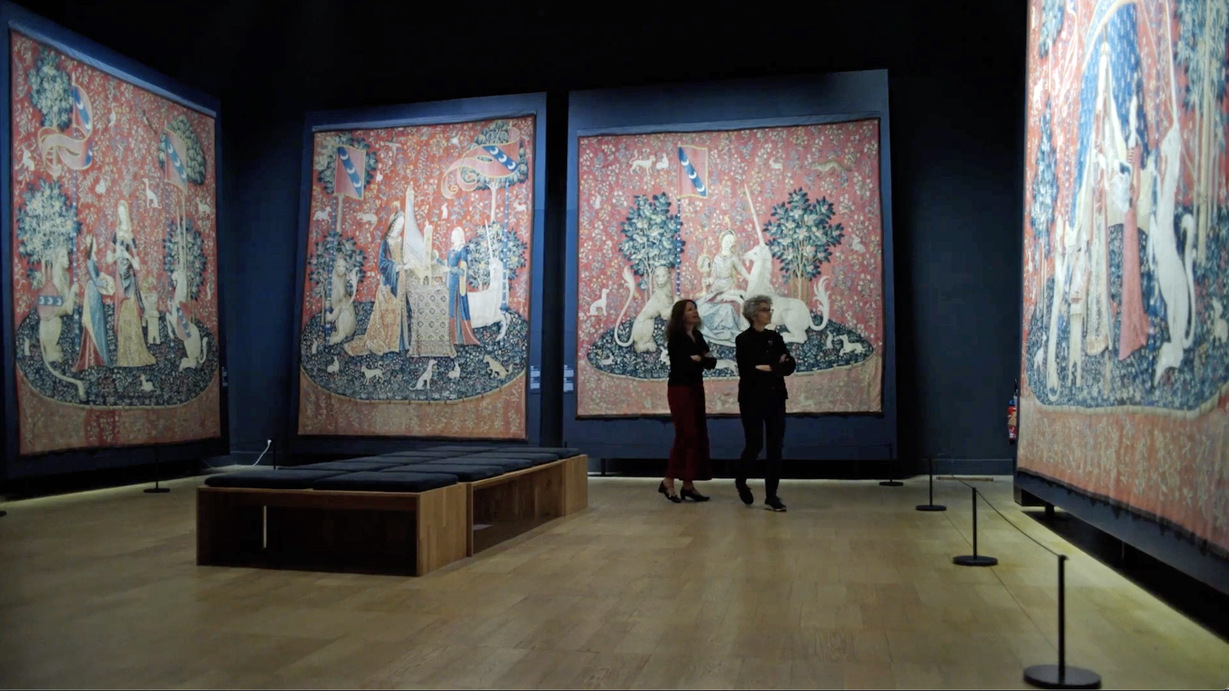 Zwei Frauen stehen vor großen, historischen Wandteppichen in einem Museum, die Szenen mit Menschen und Tieren zeigen.