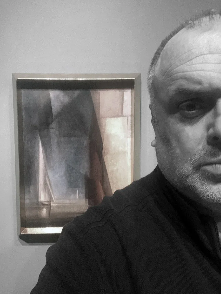 Ein Mann macht ein Selfie vor einem Kunstwerk, das geometrische, abstrakte Formen in gedeckten Farbtönen zeigt.