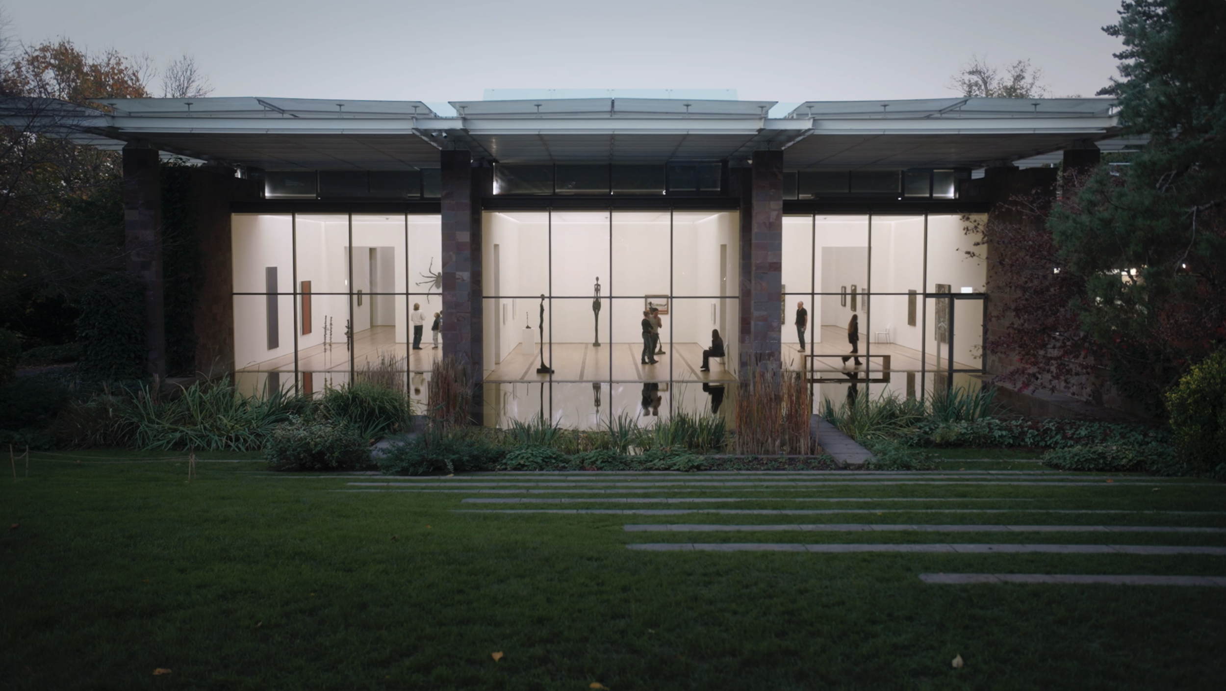 Glasgebäude mit sichtbarem Innenhof, umgeben von Bäumen, Besucher im Museum, abends, reflektierende Wasserfläche im Vordergrund.