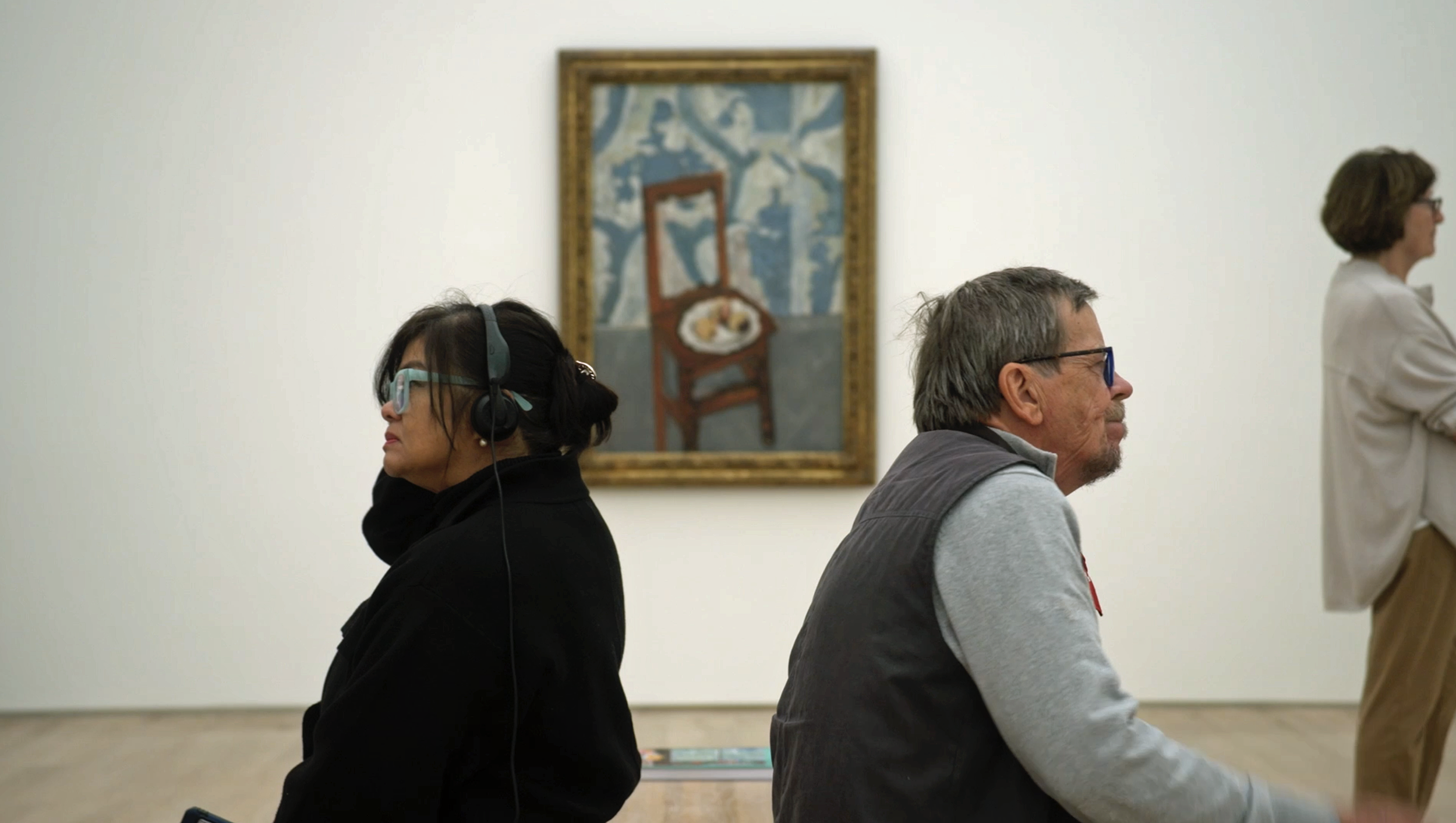 Zwei Menschen sitzen gegenüber in einem Museum, wobei eine Frau mit Kopfhörern und eine ältere Mann nebeneinander sitzen. Im Hintergrund ist ein Gemälde an der Wand.