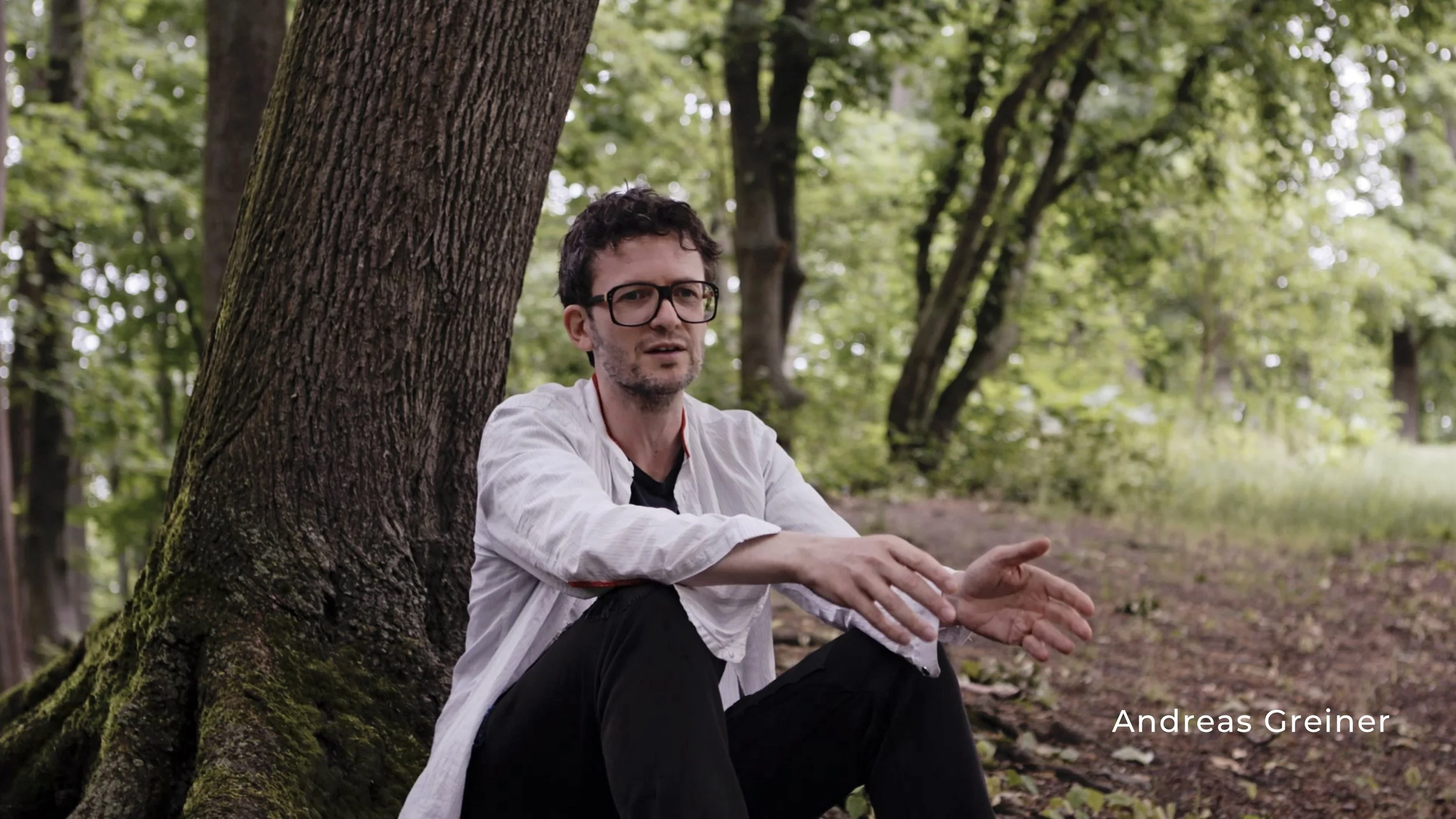 Ein Mann mit schwarzen Haaren, Brille und weißem Hemd sitzt an einem Baum im Wald.