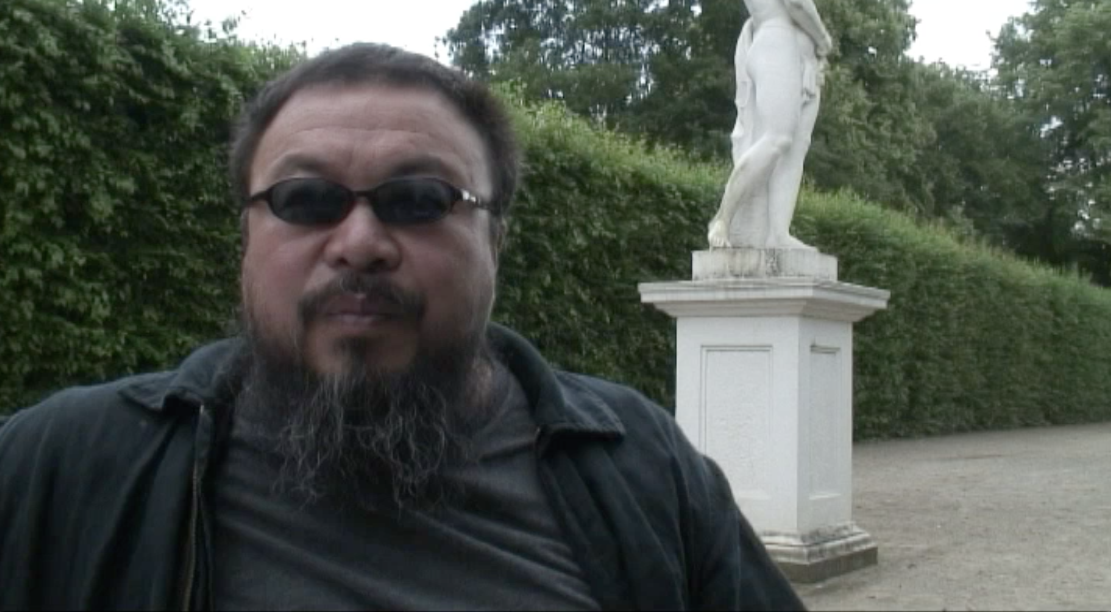 Ein Mann mit dunkler Sonnenbrille und Bart posiert vor einer weißen Statue im Freien, umgeben von Büschen und Bäumen.