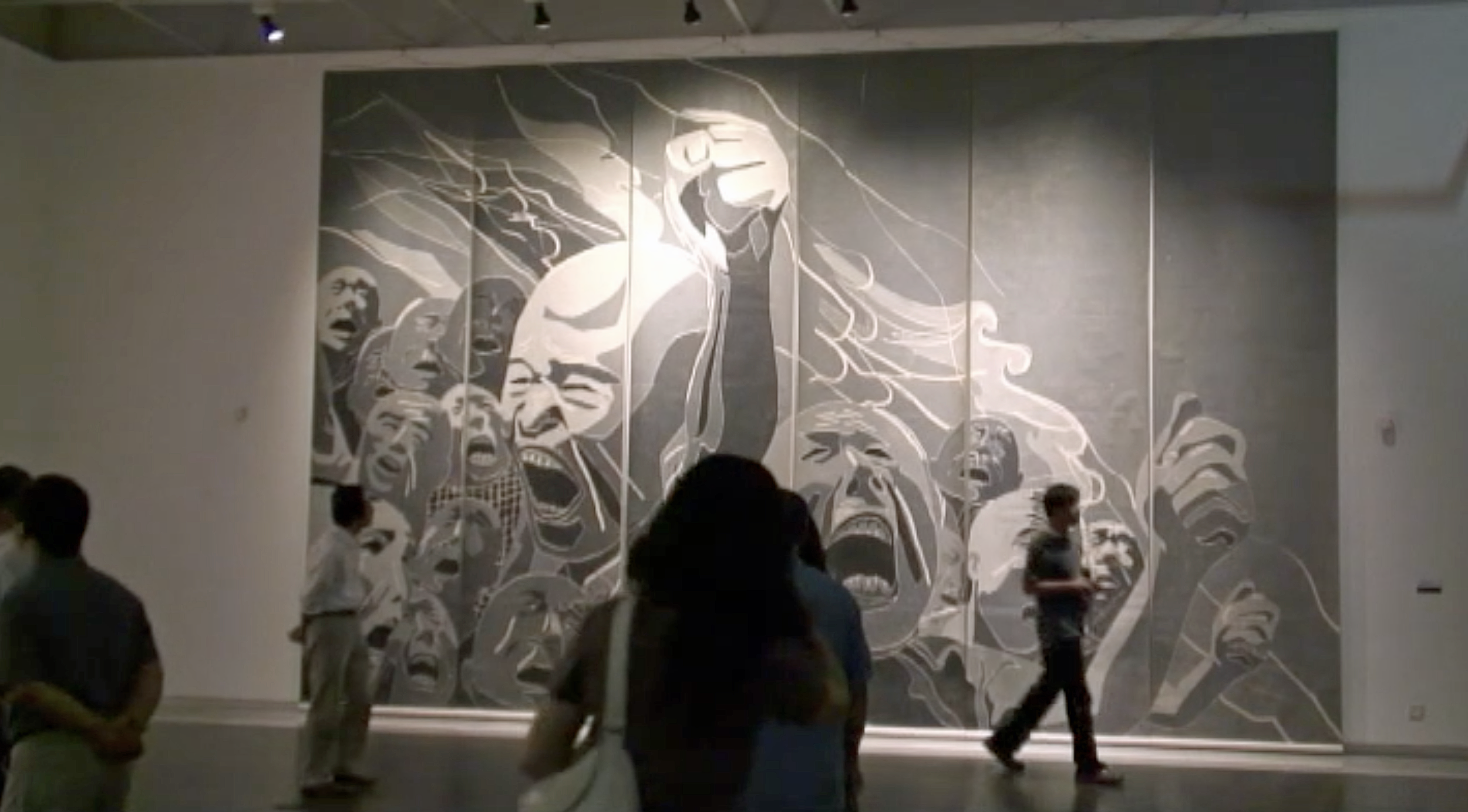 Eine große Wand mit einem schwarzen und weißen Wandbild, das mehrere ausdrucksstarke Gesichter zeigt, in einem Museum, während Menschen vorbeigehen.
