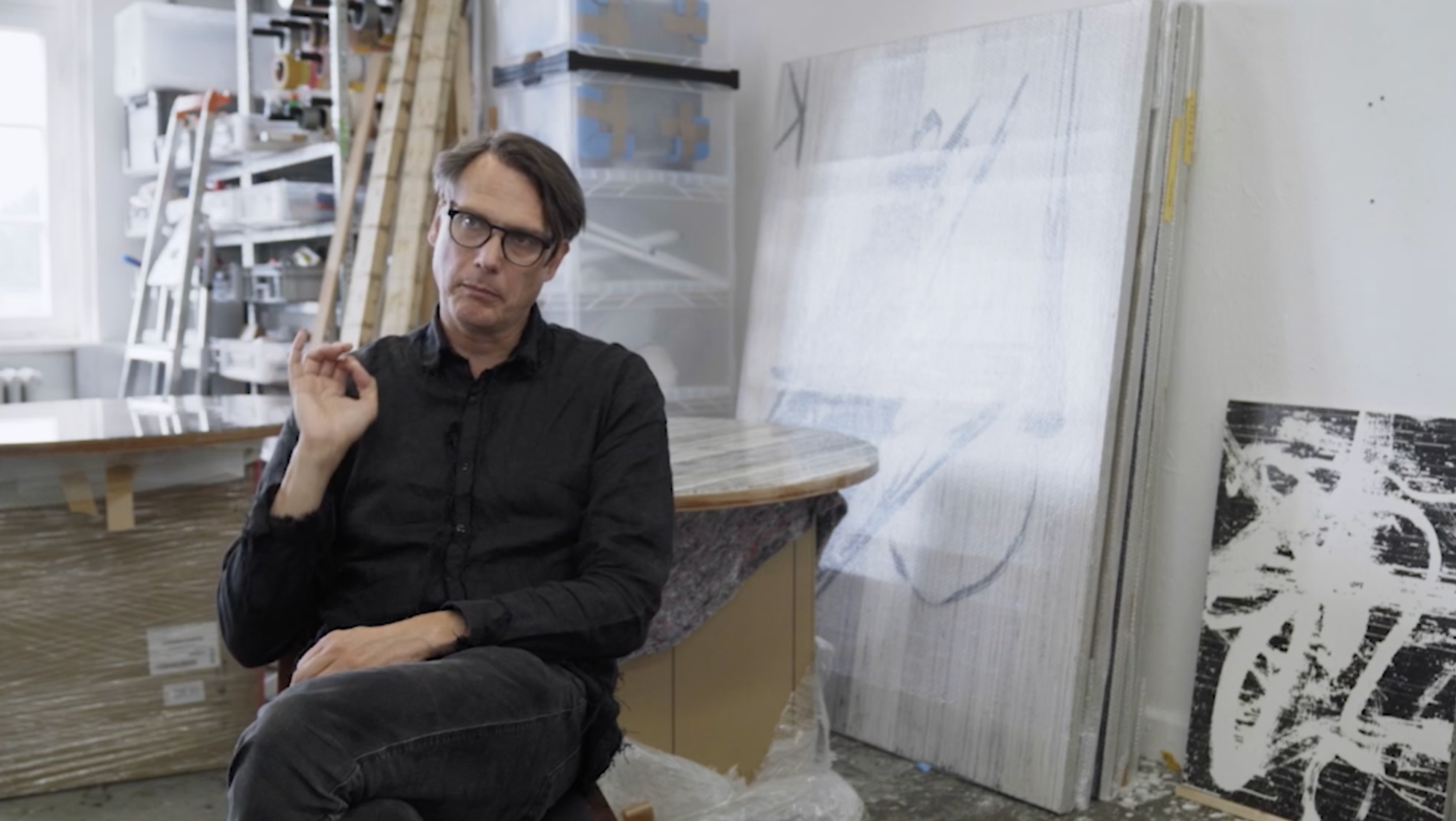 Ein Mann mit Brille sitzt in einem Kunststudio, umgeben von Leinwänden, Holzstöcken und anderen Kunstmaterialien.