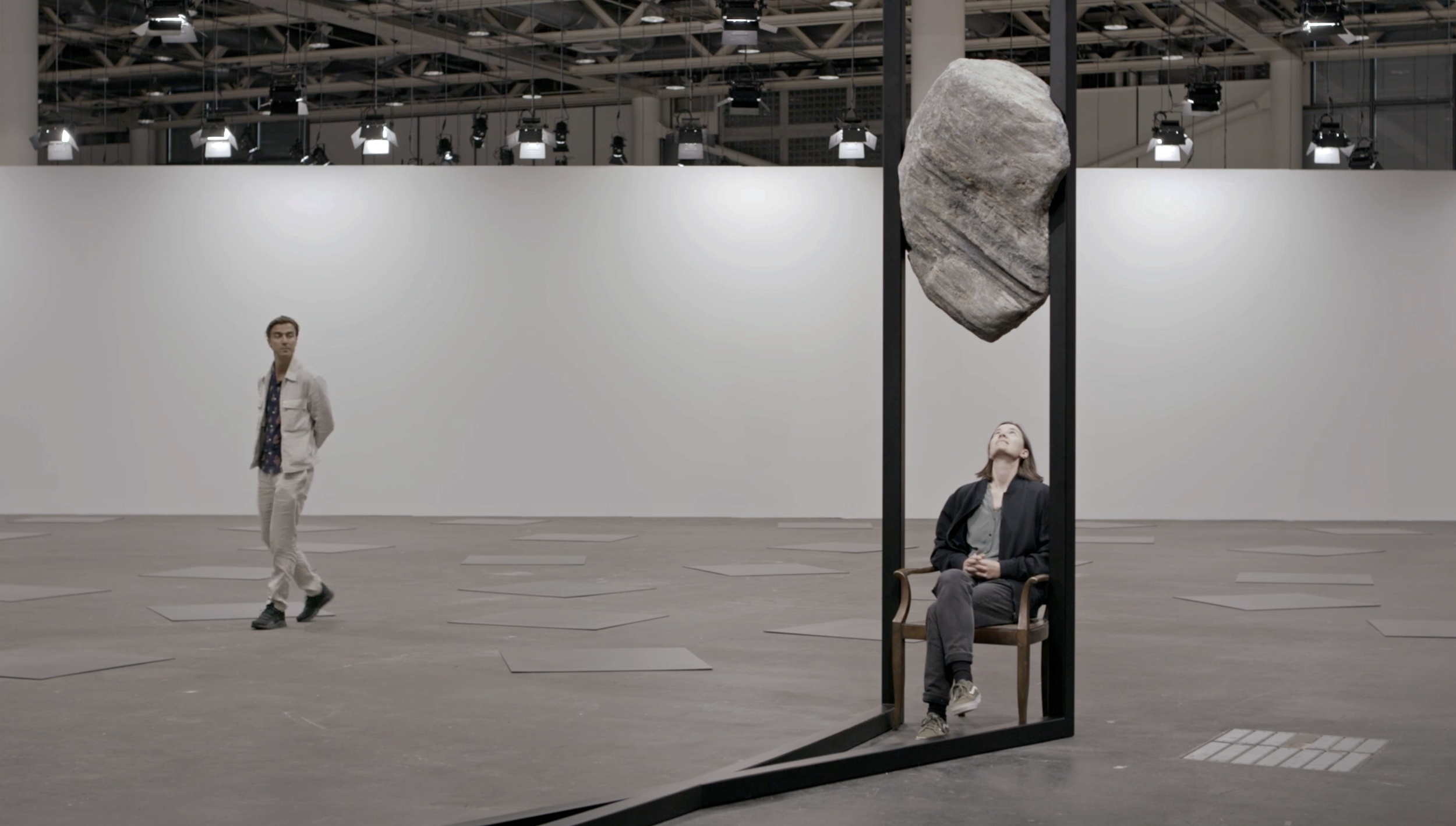 Zwei Menschen in einem modernen Kunstmuseum. Eine Frau sitzt auf einem Stuhl unter einer großen, schwebenden Steininstallation, während ein Mann in hellen Kleidern durch den Raum geht, umgeben von quadratischen Bodenplatten.