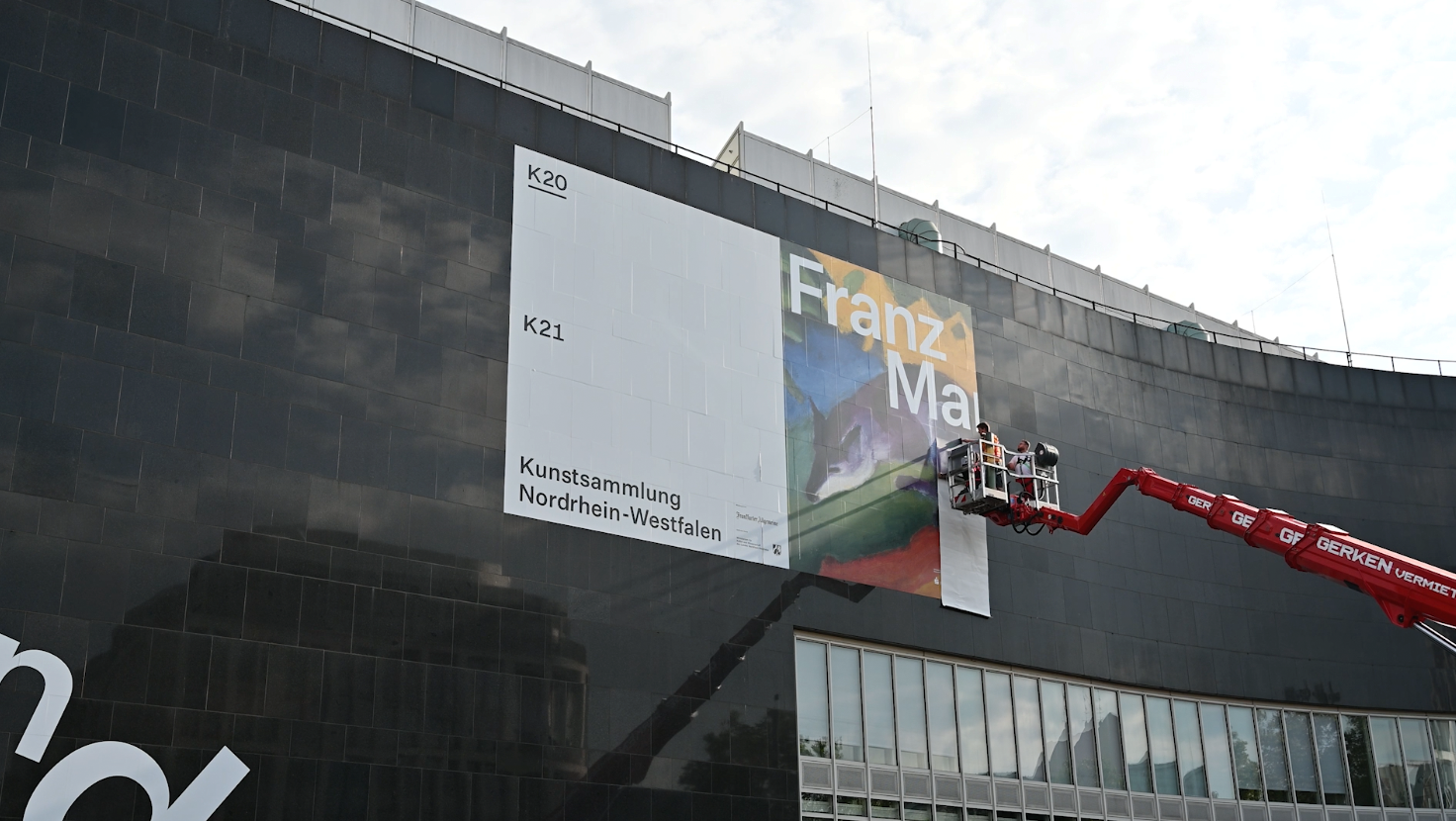 Aufbau eines Posters für die Kunstsammlung Nordrhein-Westfalen an einer schwarzen Gebäudefassade, mit einem Kran, der die Installation anbringt.